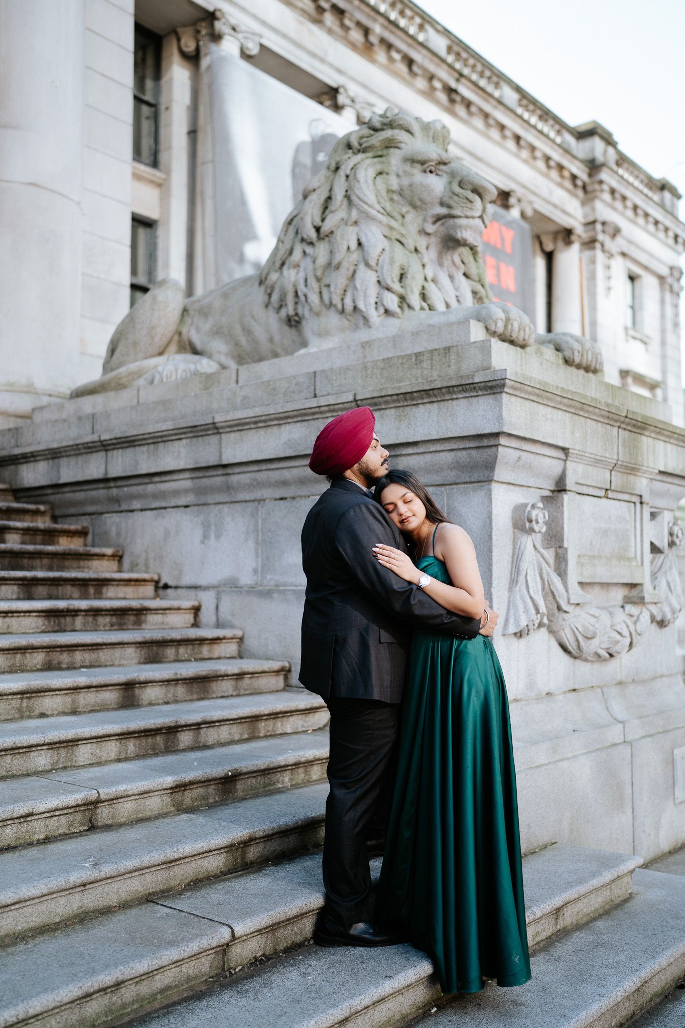 A romantic couple embraces on grand stone steps with decorative lion statues in an elegant architectural setting.