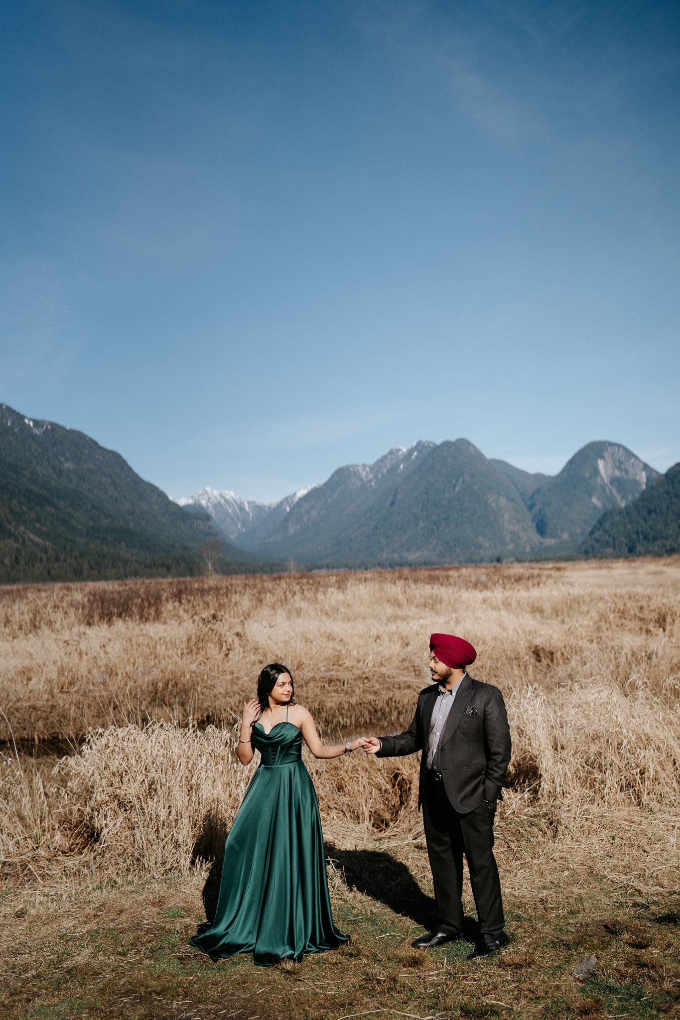 A couple in formal attire poses in a scenic mountain meadow with snow-capped peaks in the background.