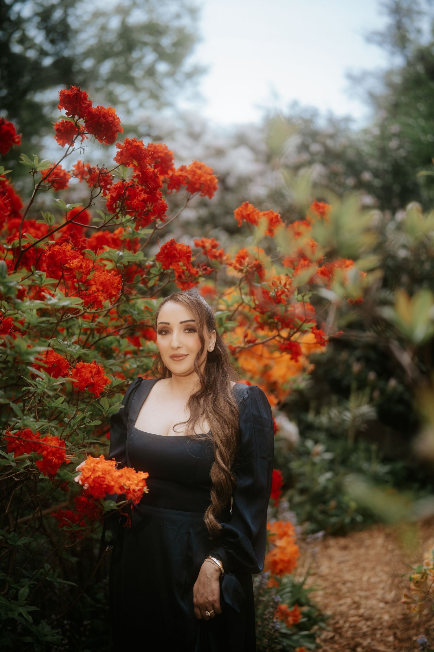 Woman in black dress posing among vibrant red and orange flowering bushes in a lush garden setting.