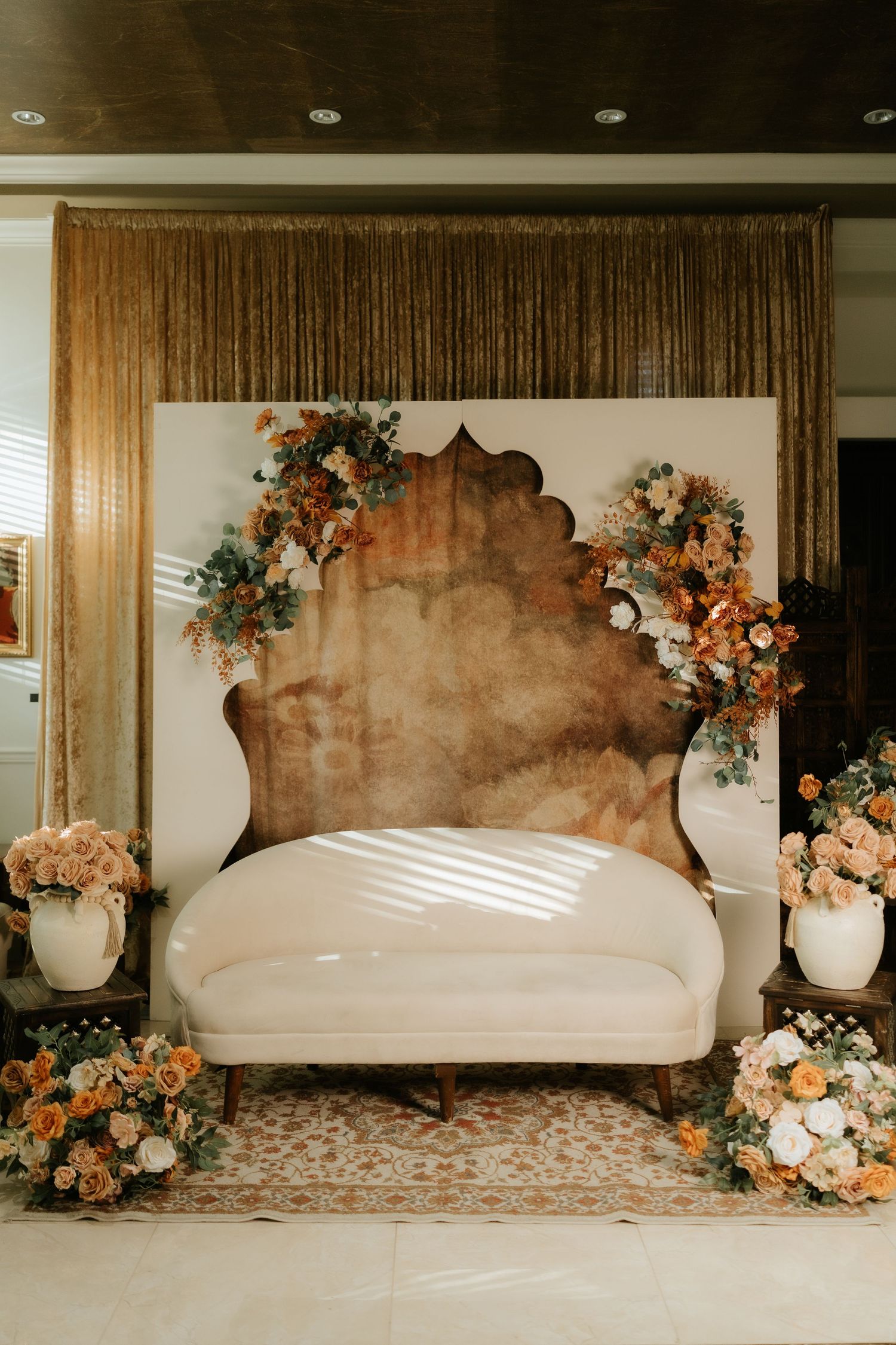 Elegant wedding backdrop with white loveseat, rust floral arch, peach roses, and gold sequin curtains for a romantic ceremony.