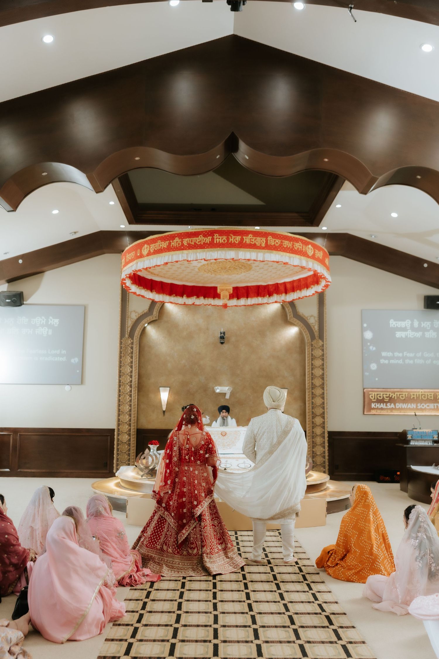 Sikh wedding ceremony with bride in red lehenga and groom in white at Gurdwara altar under decorative canopy.