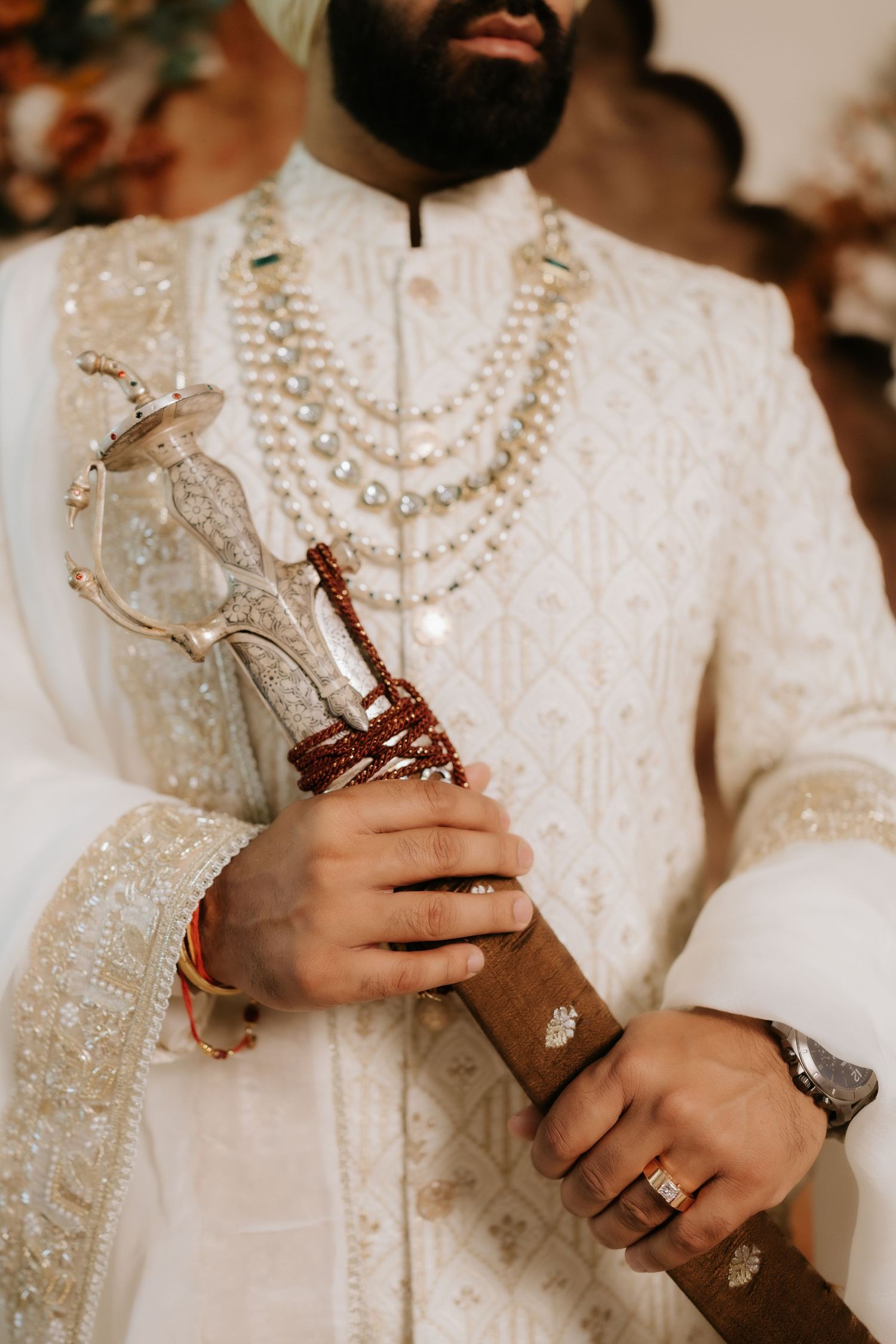 Sikh groom in white embroidered sherwani with pearl necklaces holding a ceremonial kirpan at a wedding.