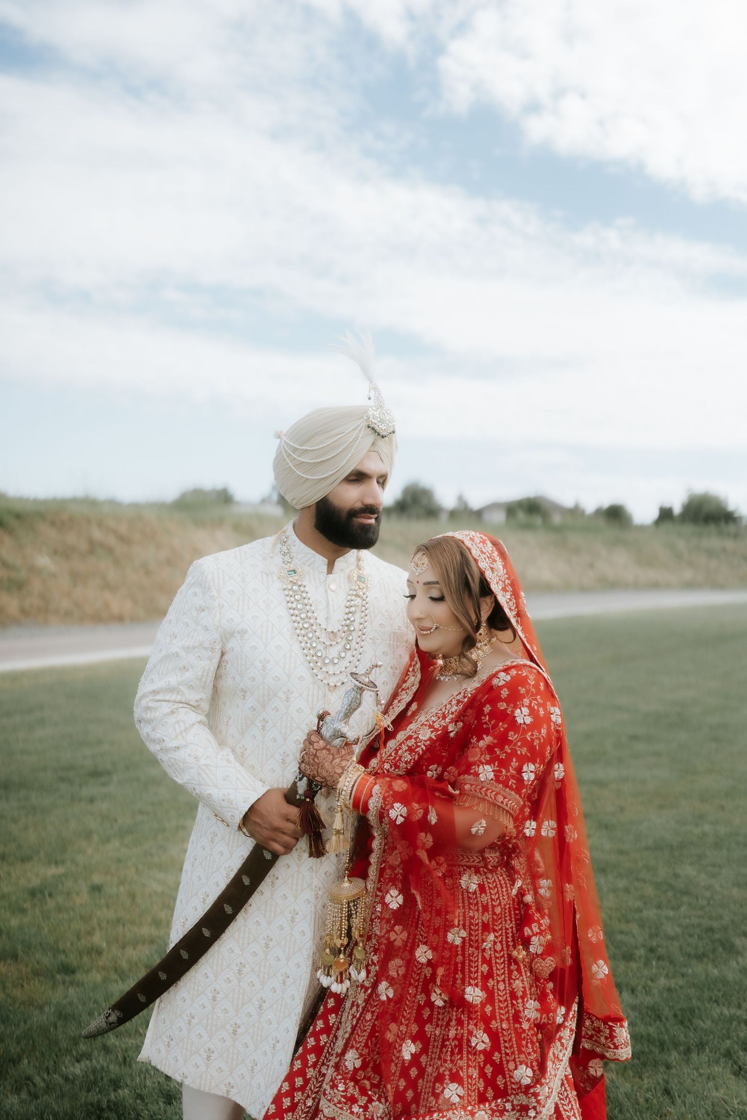 Sikh bride in red lehenga and groom in white sherwani with turban pose outdoors on their wedding day.