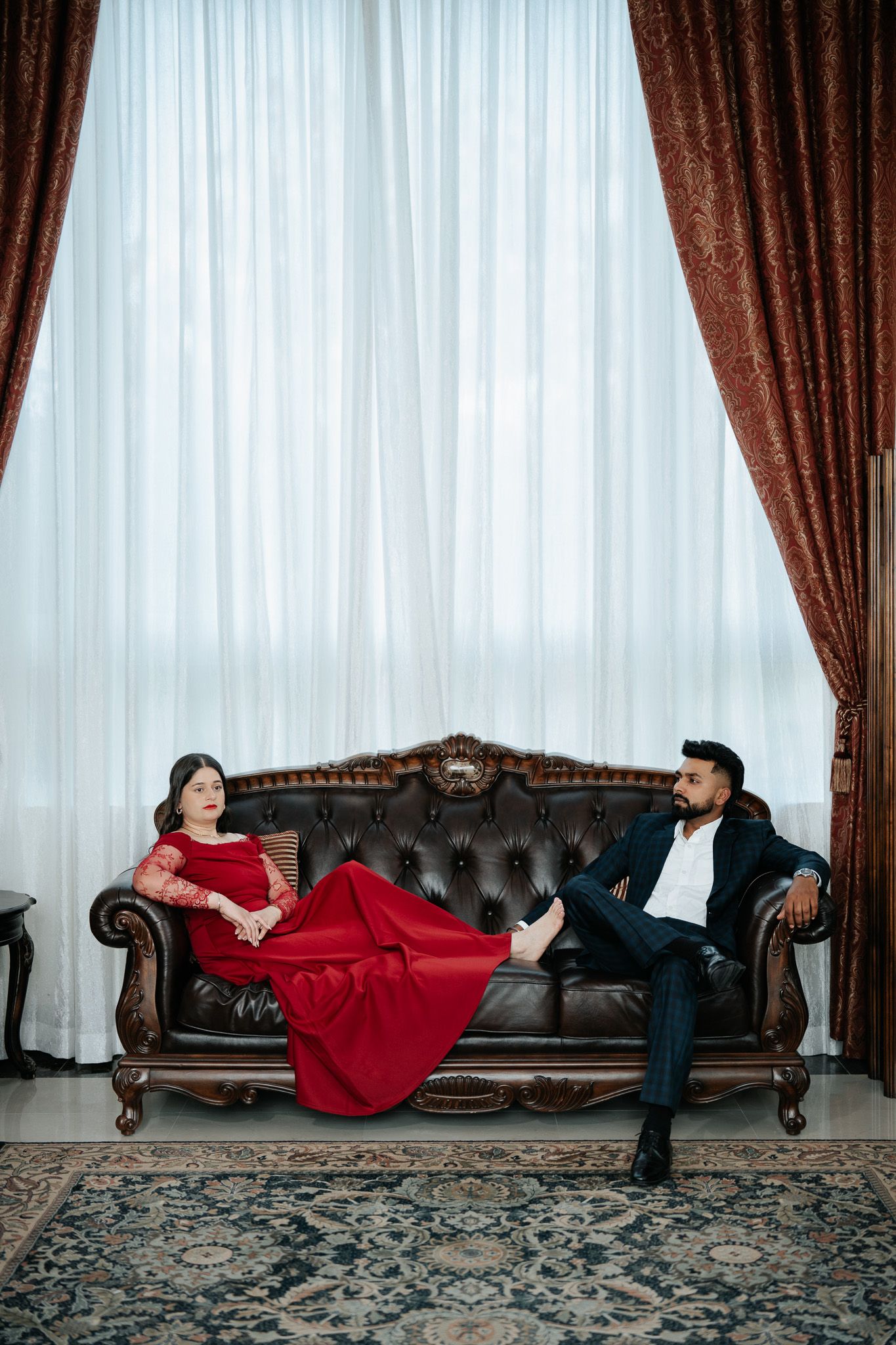 Elegant couple on a vintage leather sofa, woman in red gown and man in suit, posed before sheer curtains.