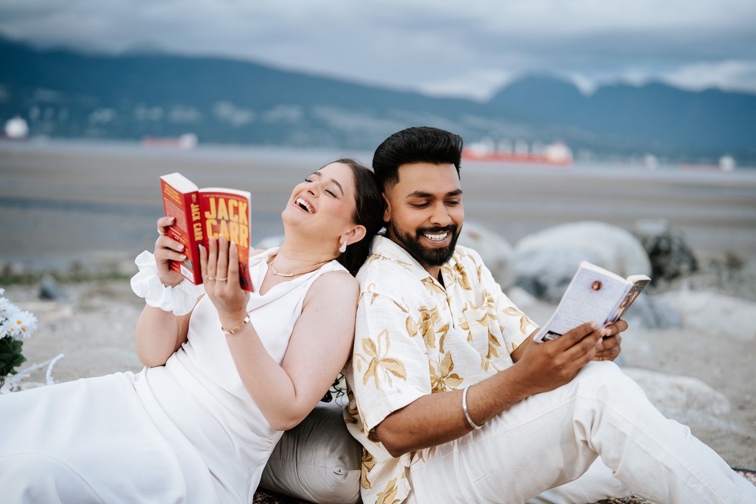 Laughing couple sitting back-to-back outdoors, woman holding red book and man smiling at tablet, scenic mountain backdrop.