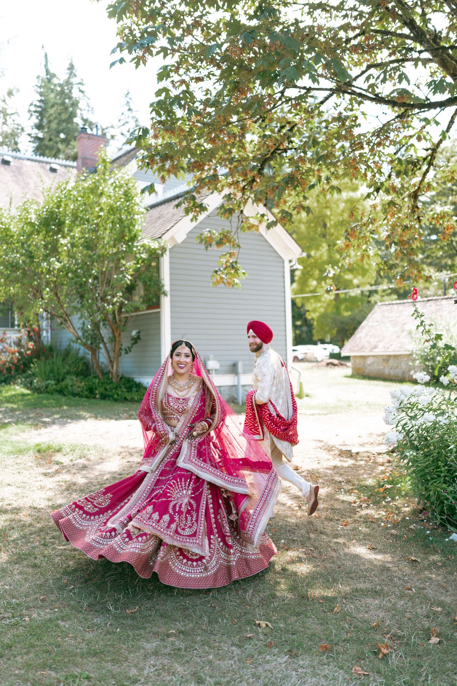 Sikh bride in magenta lehenga twirls outdoors as groom in red turban walks behind during wedding celebration.