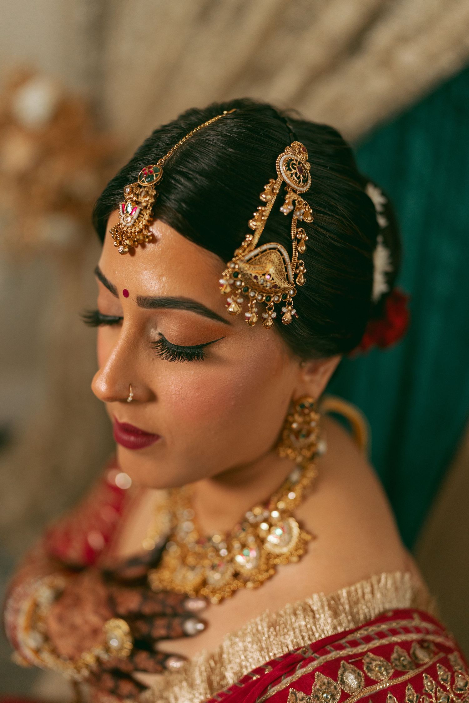 Indian bride in red saree wearing gold maang tikka, nose ring, and layered necklace with mehndi hands.