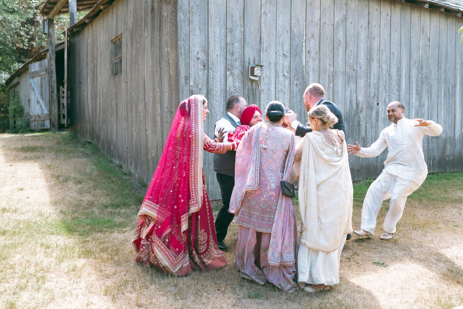 South Asian wedding party in traditional attire dancing outdoors near a rustic wooden barn on a sunny day.