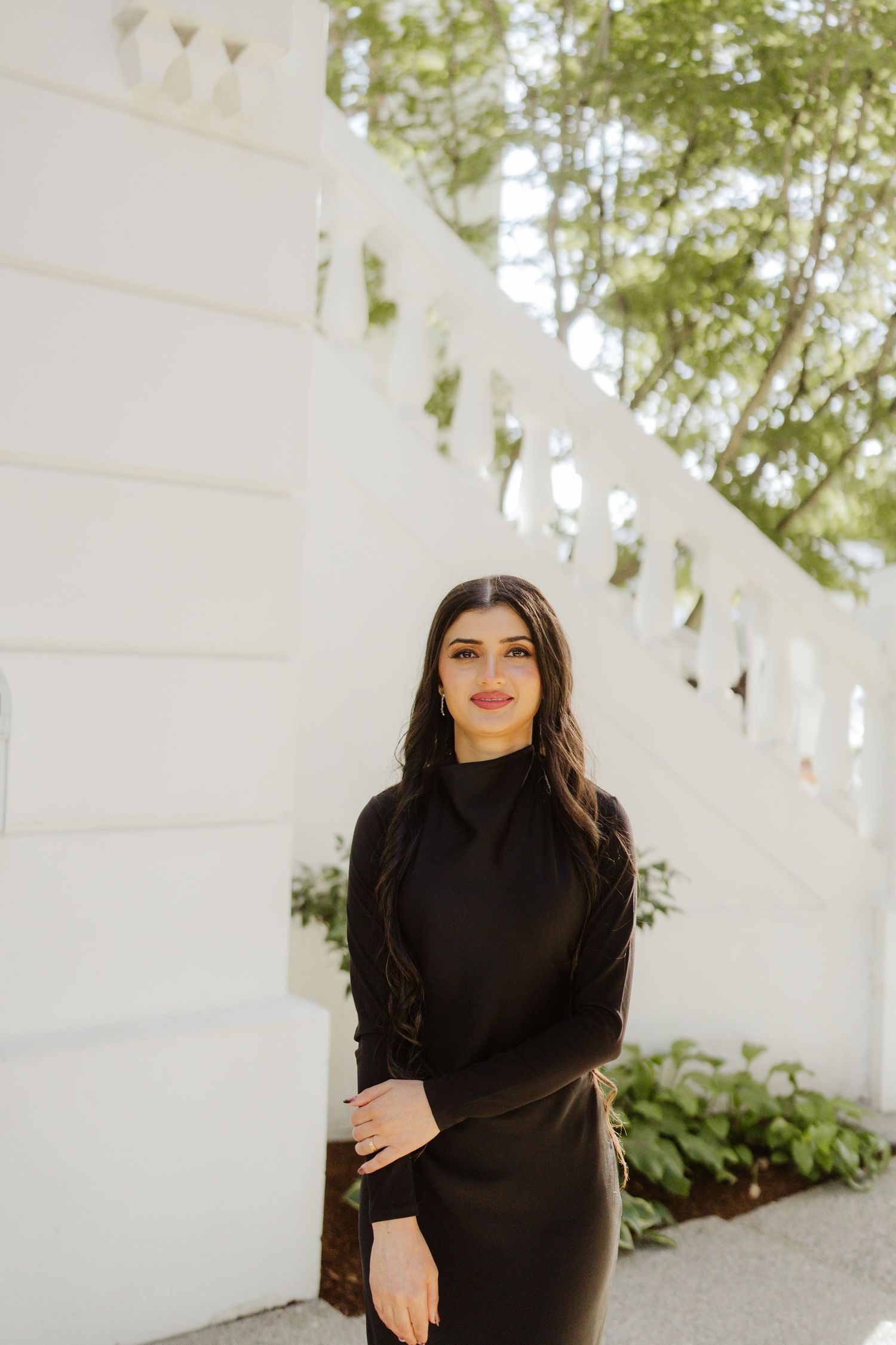 Young woman with long dark hair wearing a black turtleneck dress posing by a white wall.