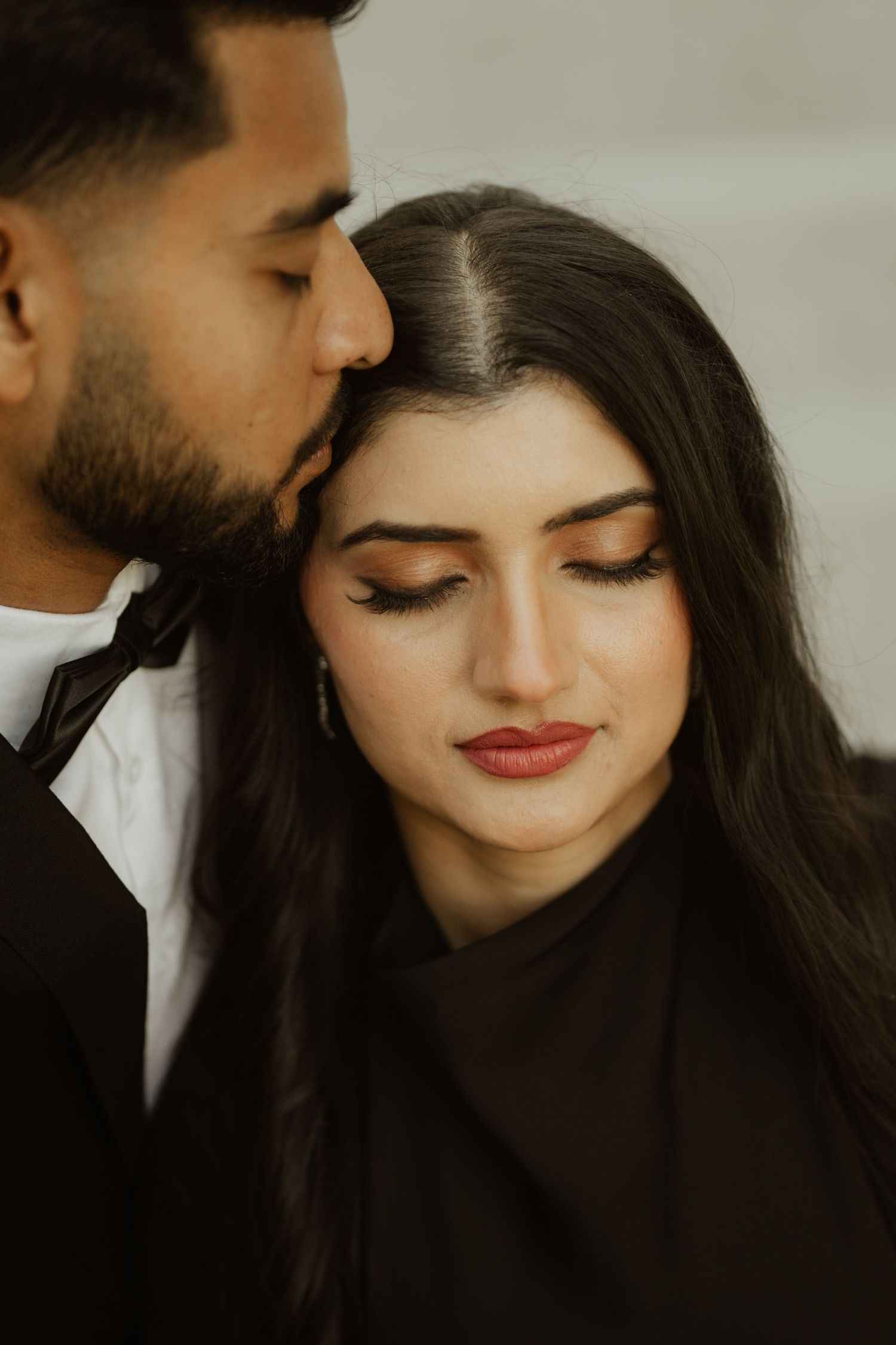 A man tenderly kisses a woman on the forehead as she closes her eyes, both dressed in black.