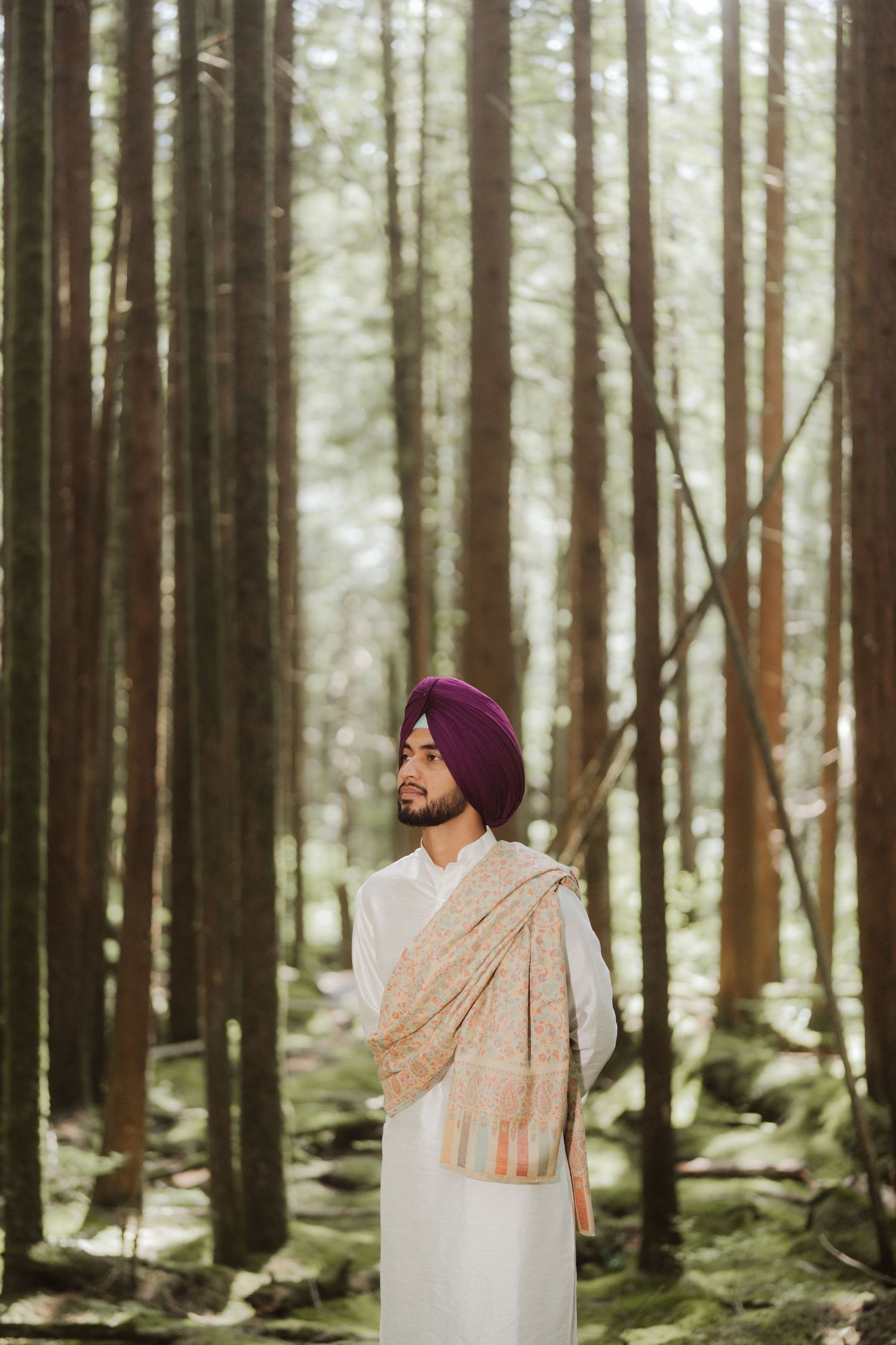 Sikh man in purple turban and white kurta with pashmina shawl standing in a sunlit forest.