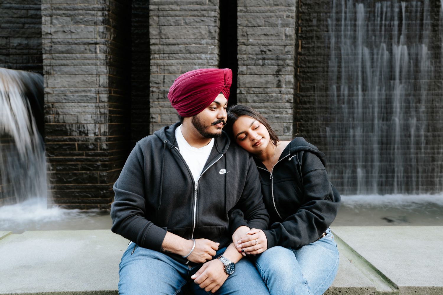 A couple sits together on stone steps near a cascading waterfall in an urban setting.