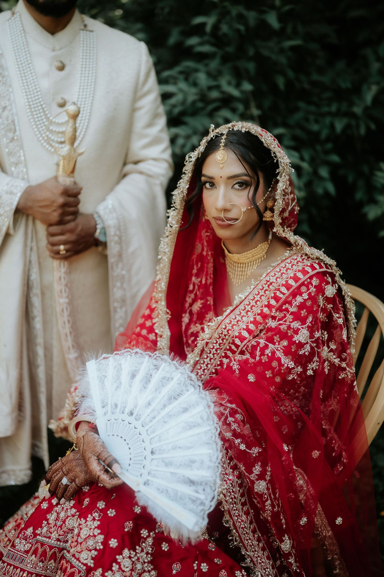 A wedding couple in traditional Sikh attire poses together with the bride holding a white decorative fan.