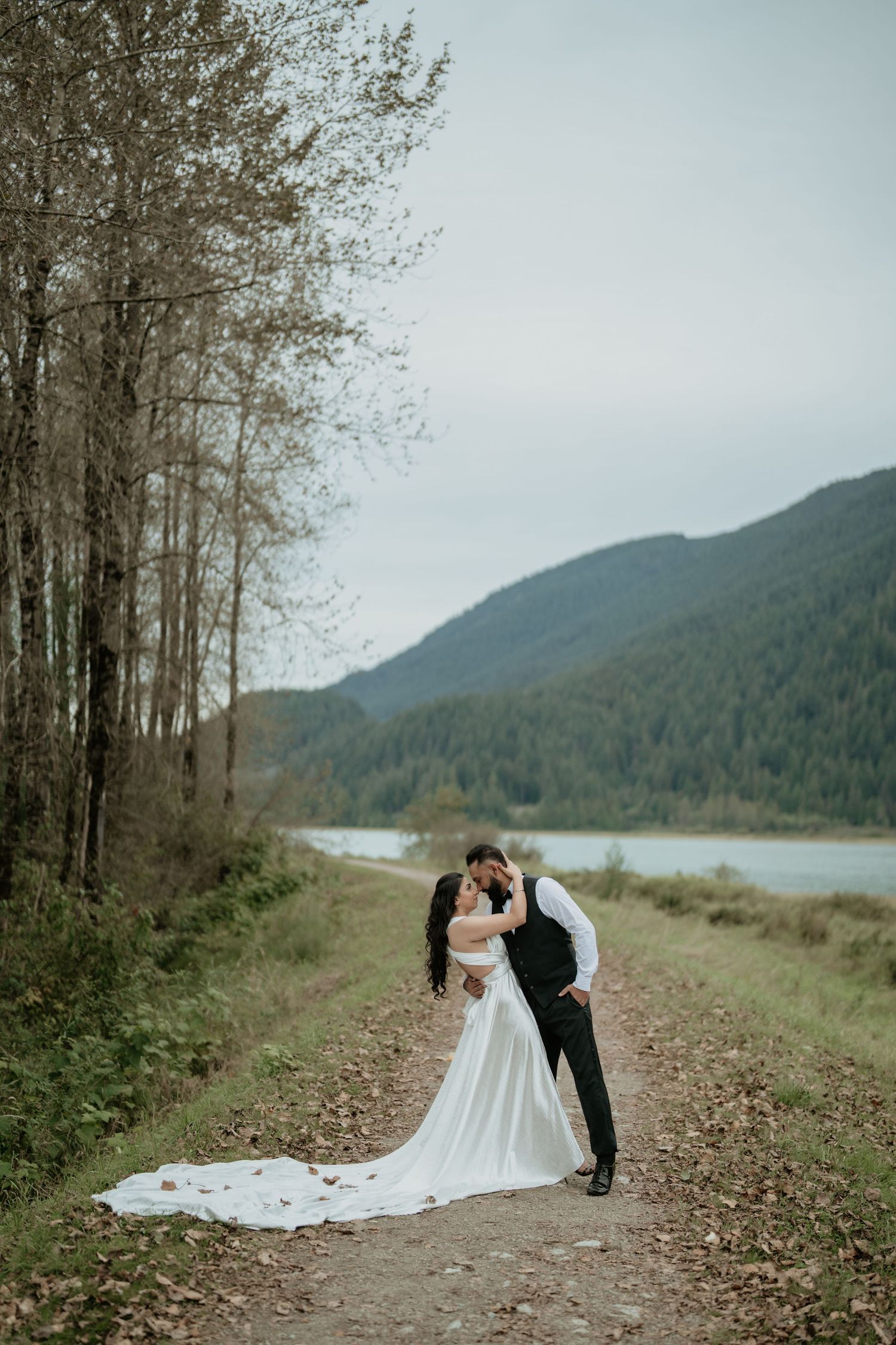 Wedding couple poses along scenic mountain lake trail with long flowing dress.