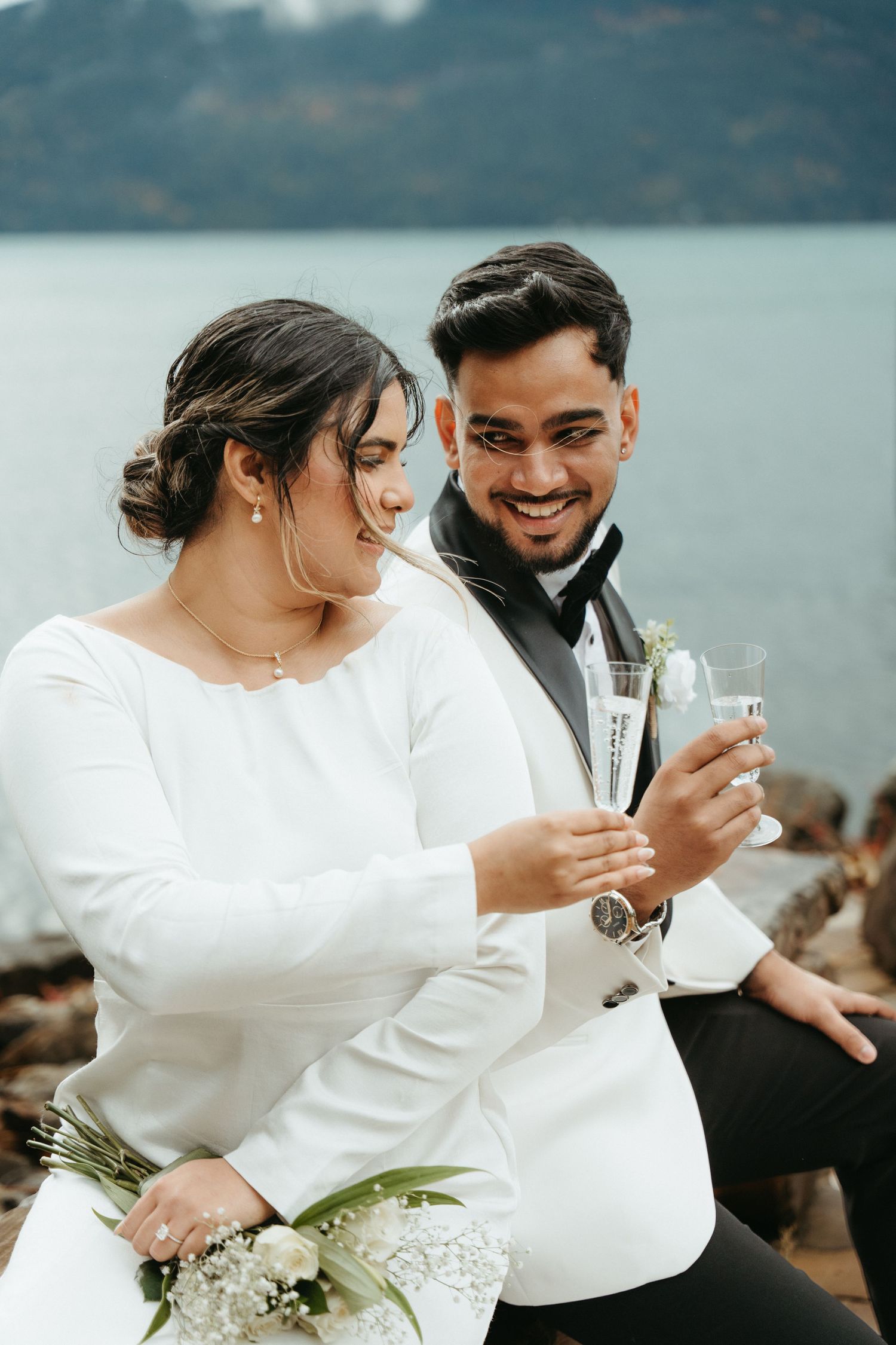 A couple in white outfits share a romantic moment while looking at their phones by a scenic waterfront.