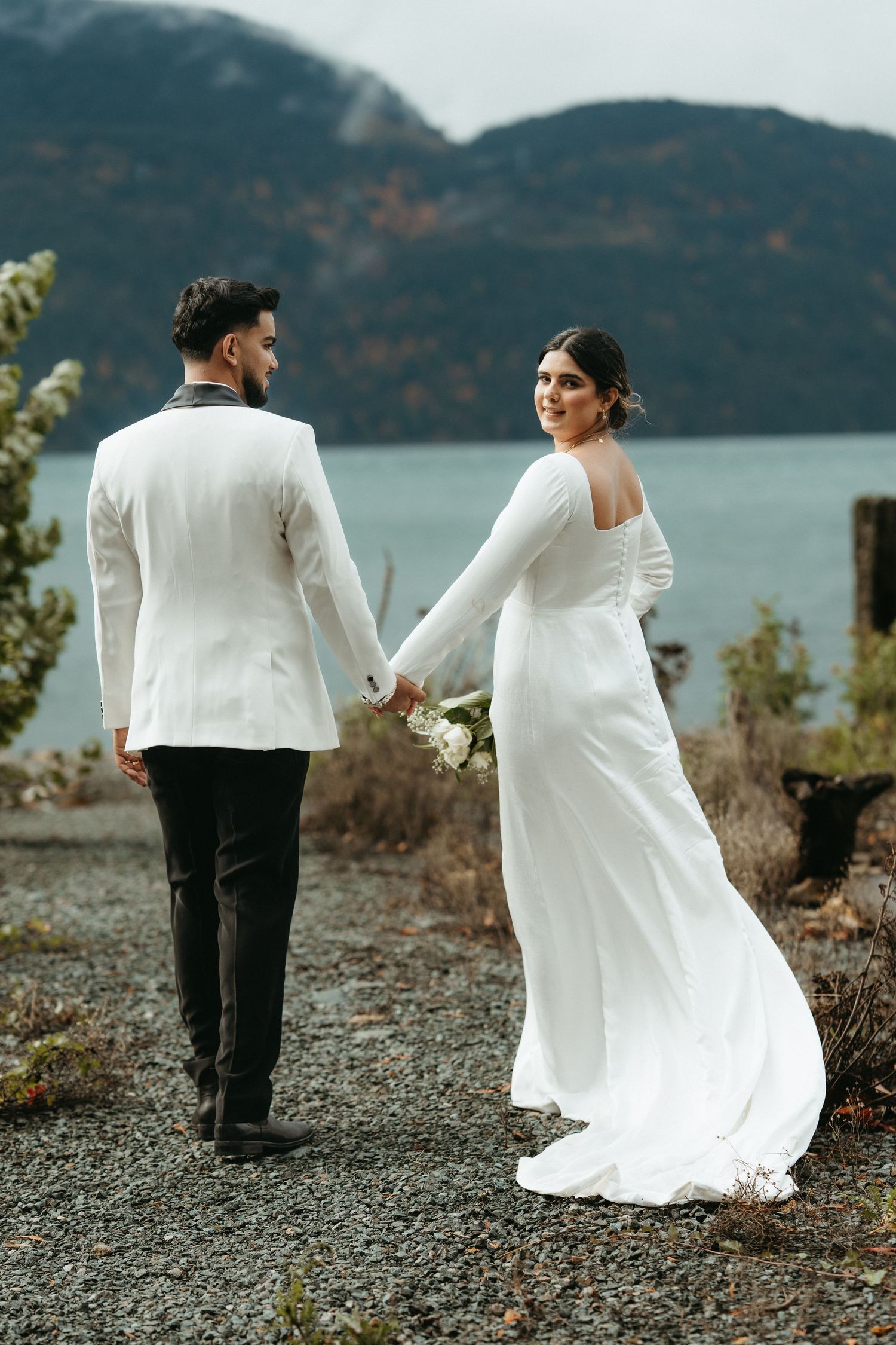 A couple in wedding attire walks along a scenic mountain lakeside path with flowing white dress and dark suit.