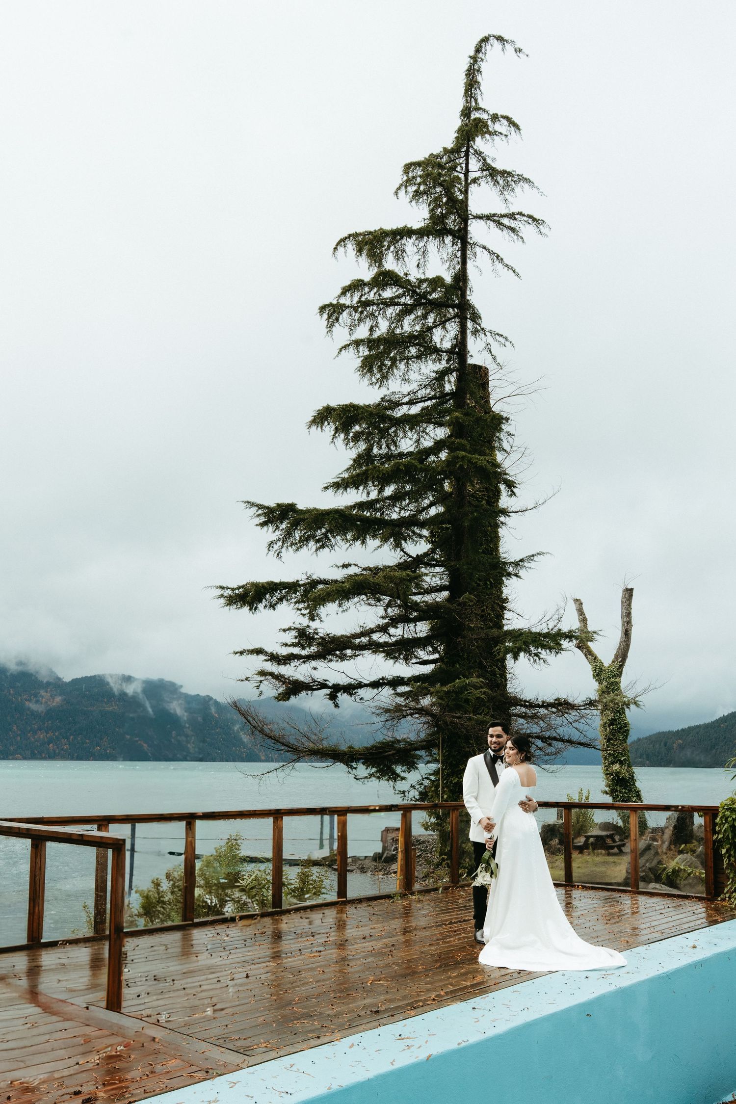 Someone in a white wedding dress stands on a deck overlooking a misty mountain lake with a tall evergreen tree nearby.
