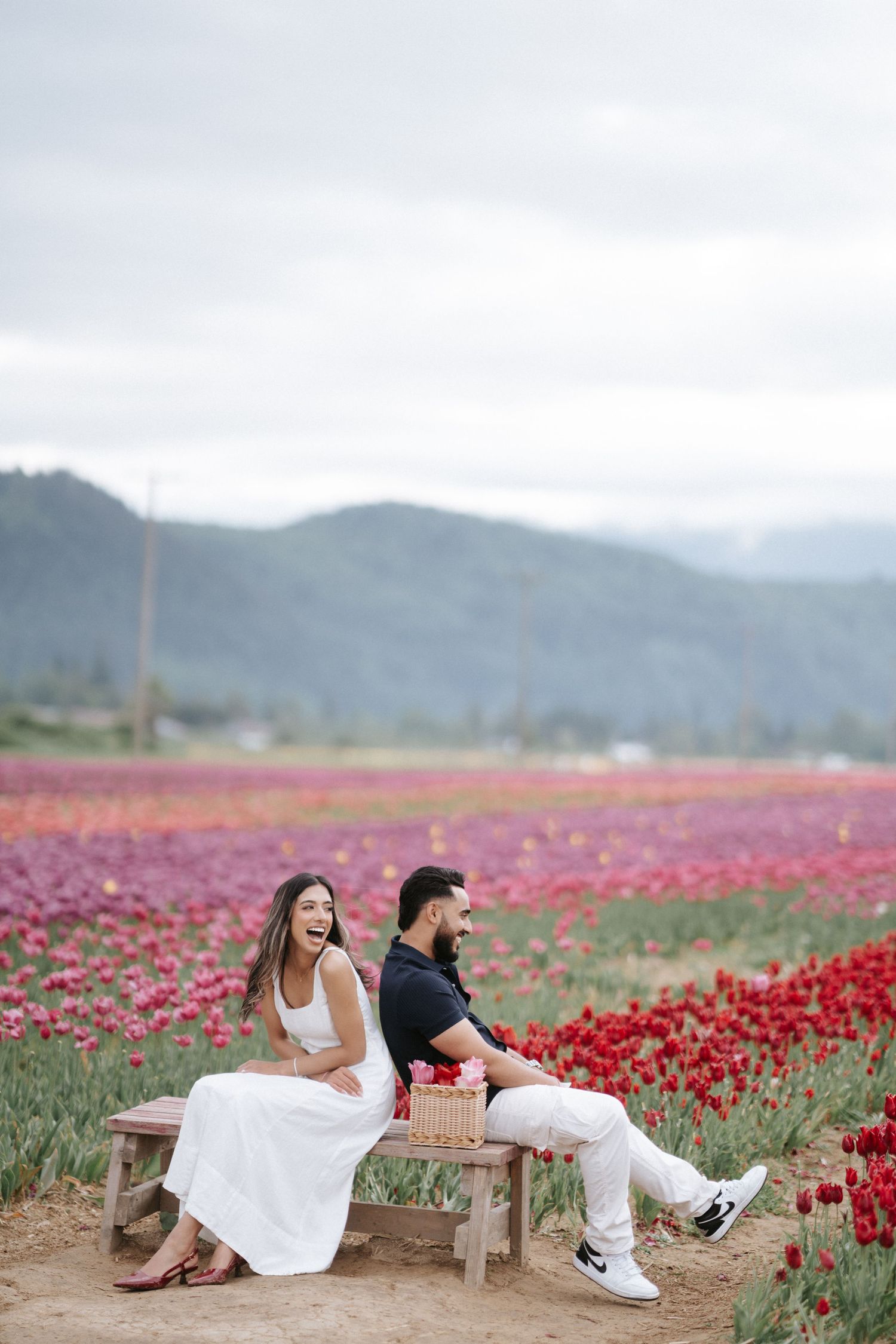 Two people sit back-to-back on wooden benches surrounded by vibrant red tulip fields with mountains in the distance.