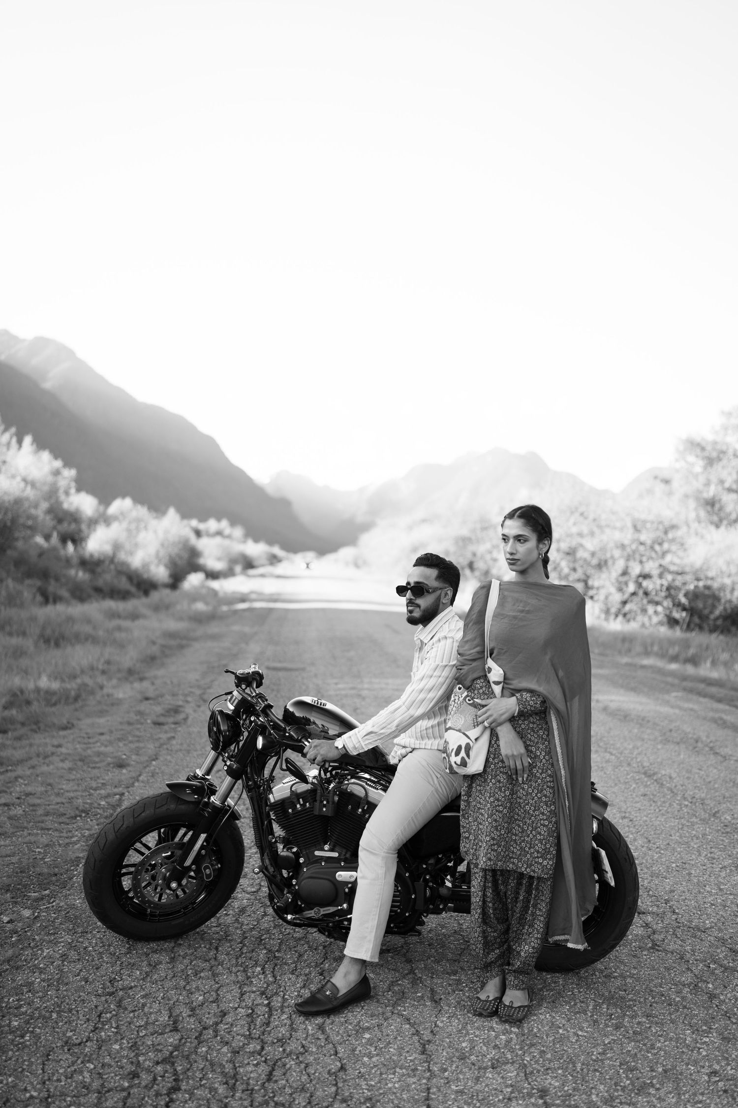 A couple in vintage attire relaxes on a classic motorcycle in black and white against a scenic mountain backdrop.