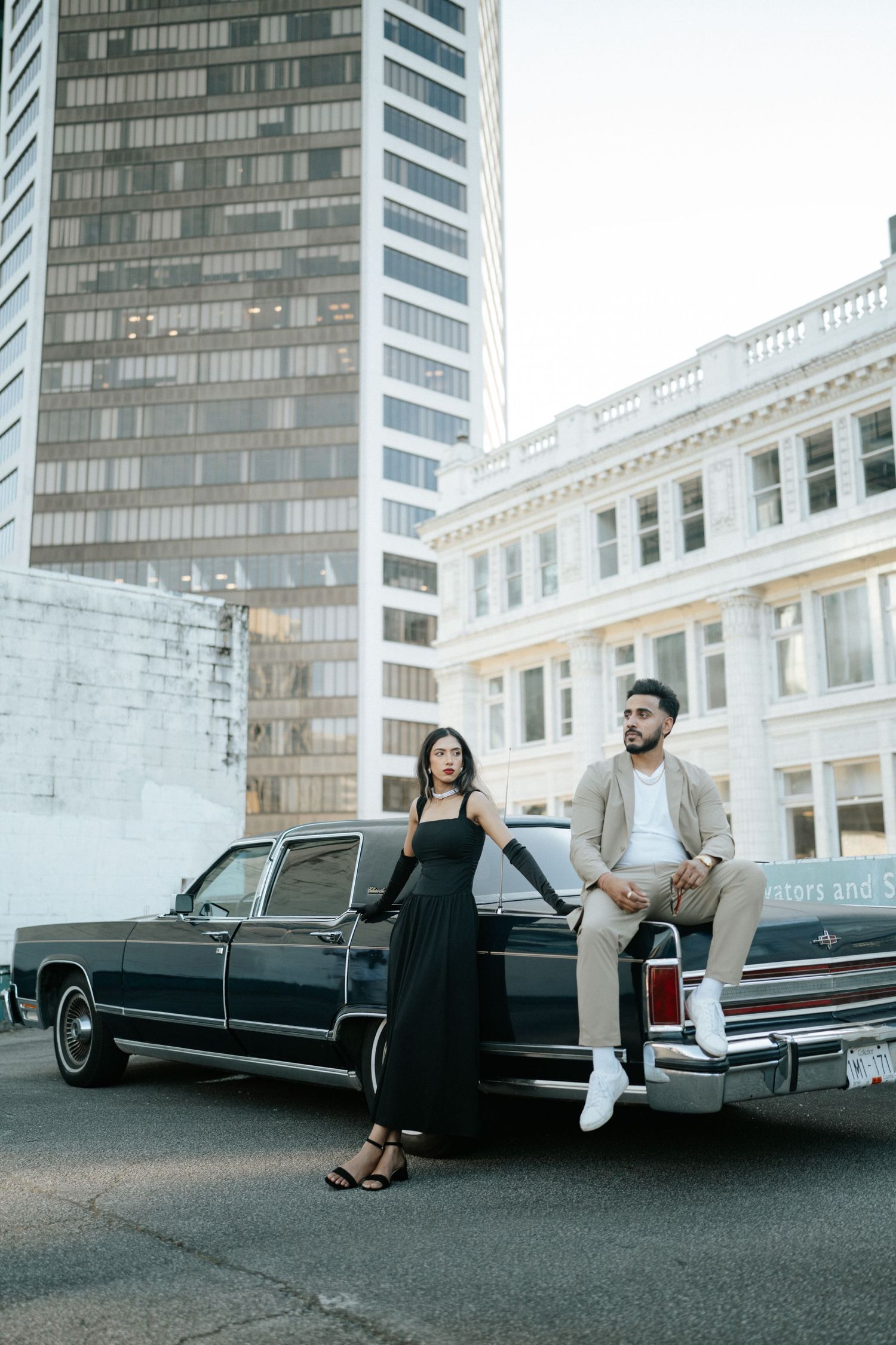 A couple poses stylishly with a vintage black limousine against an urban backdrop with modern buildings.