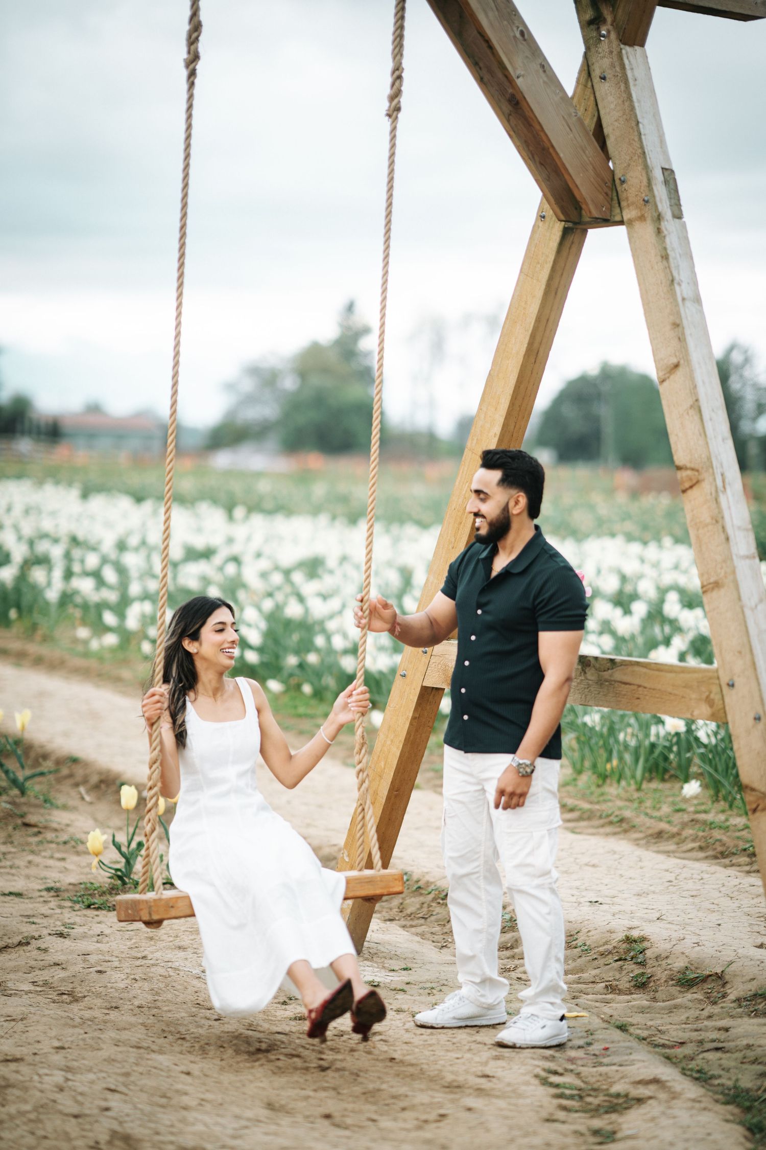 A couple in white outfits plays on a wooden swing in a field of white wildflowers on a cloudy day.