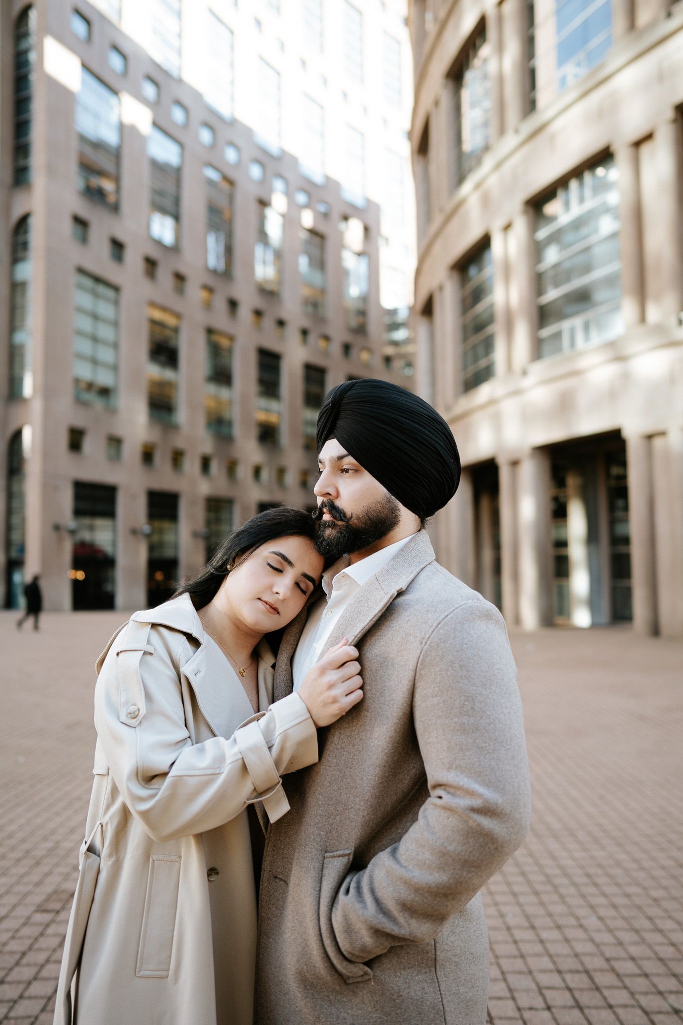 A couple in beige coats embrace while standing in front of modern urban architecture on a cobblestone plaza.