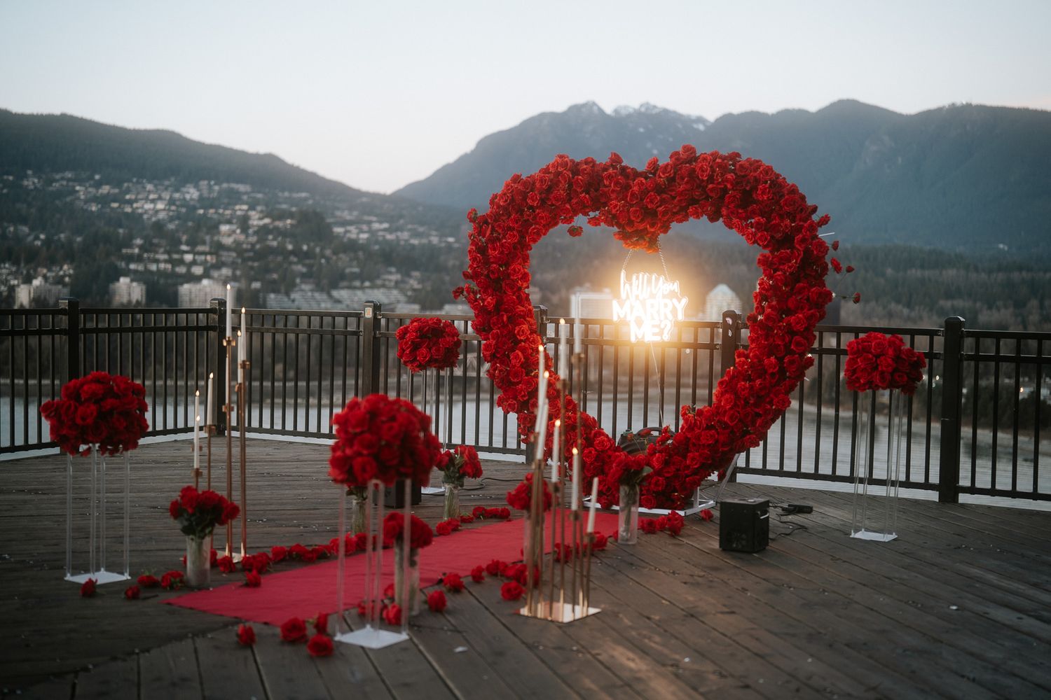 A romantic red heart-shaped wedding arch on a mountain view deck at sunset with rose decorations and red carpet.