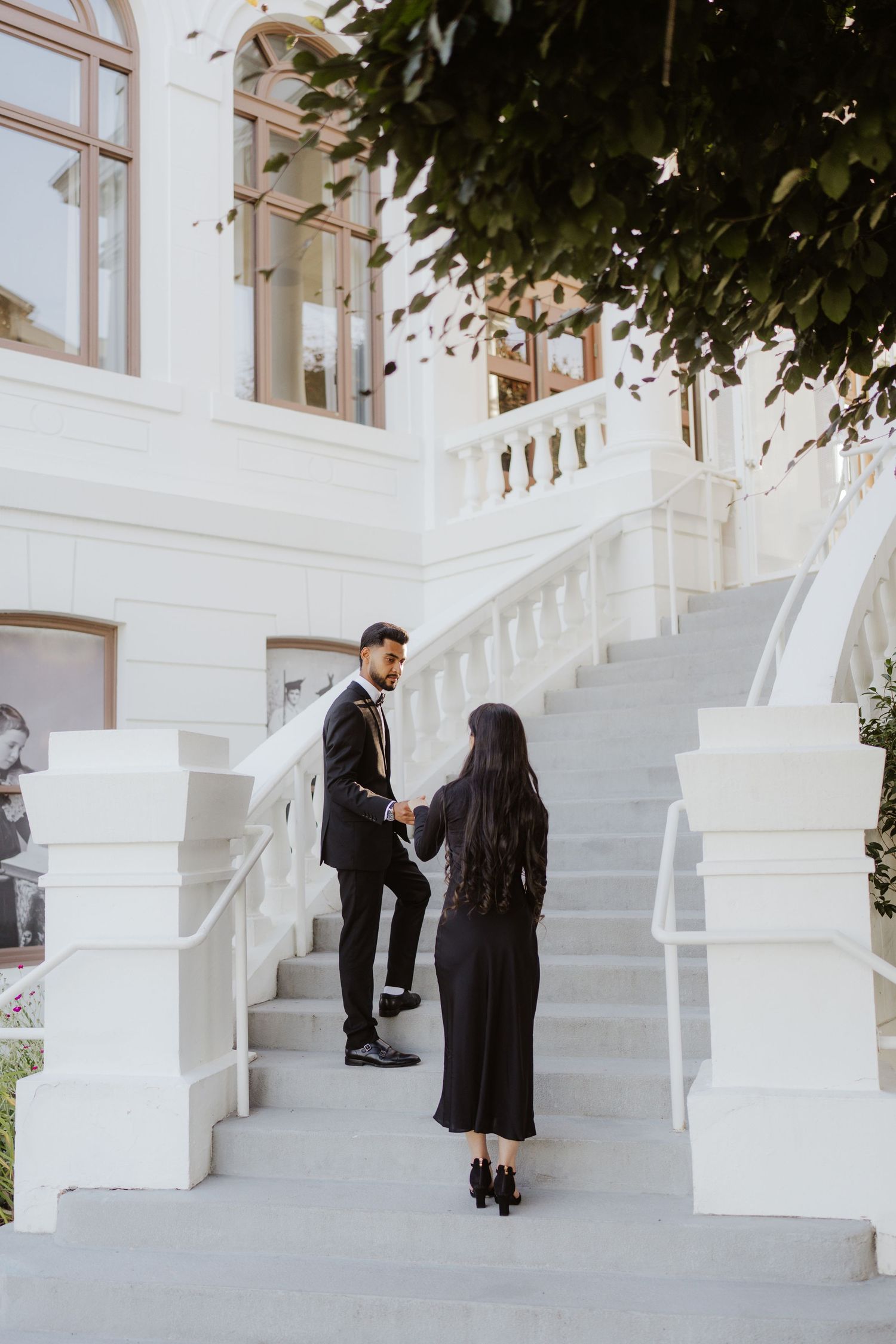 An elegant couple in formal black attire hold hands on white marble stairs outside a grand building.