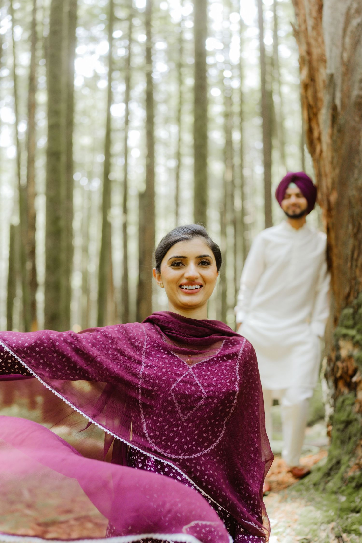 A smiling woman in a purple embroidered salwar suit poses in a forest with a Sikh man in white behind her.