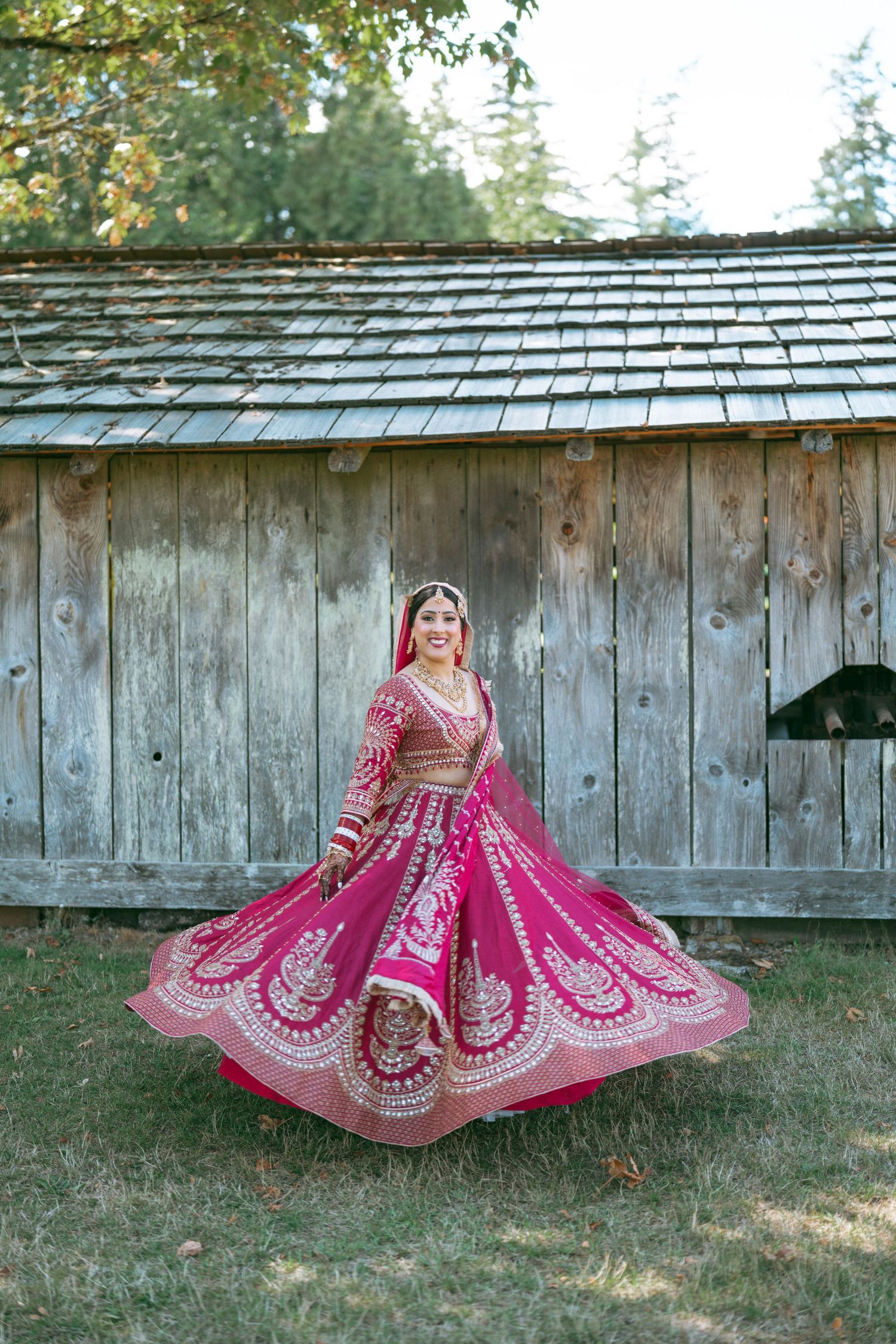 Bride in vibrant pink embroidered lehenga twirling outdoors near rustic wooden barn, showcasing traditional South Asian bridal attire.