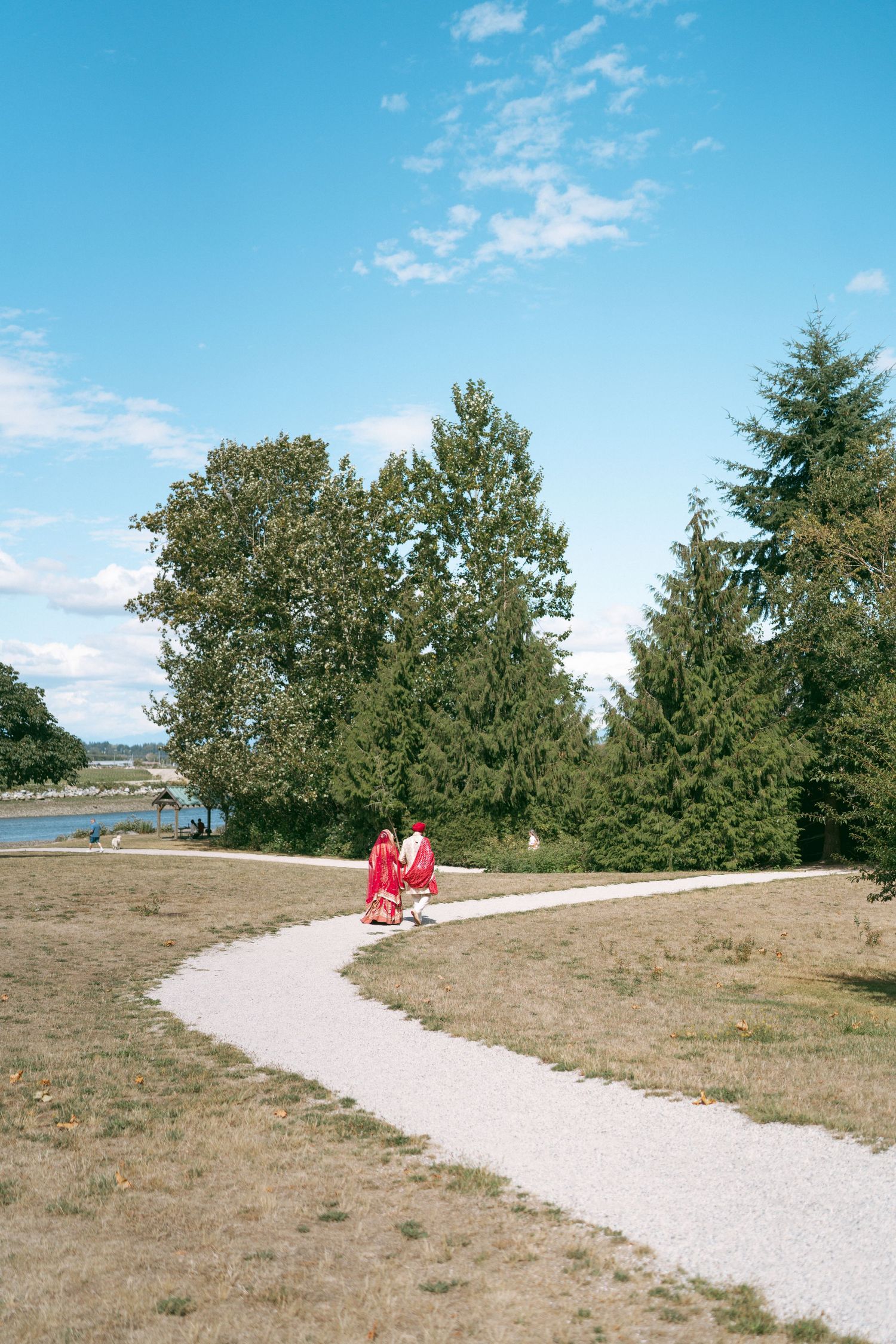 Two people in red outfits walk along a winding path in a lush green park near a waterfront under blue skies.