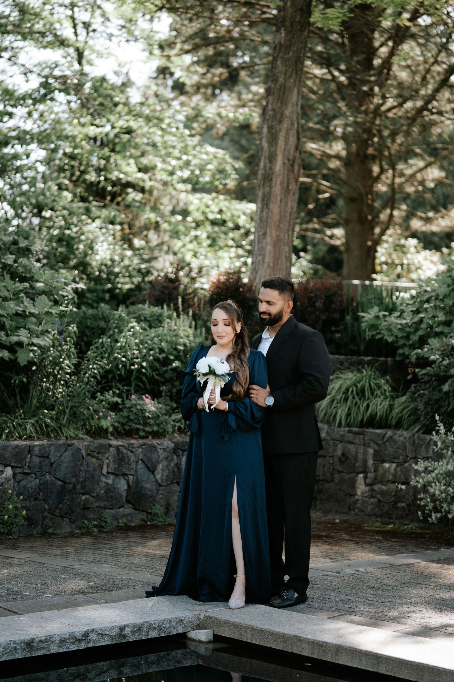 Couple in elegant dark formalwear poses in lush garden, woman holding white bouquet beside stone wall and pond.