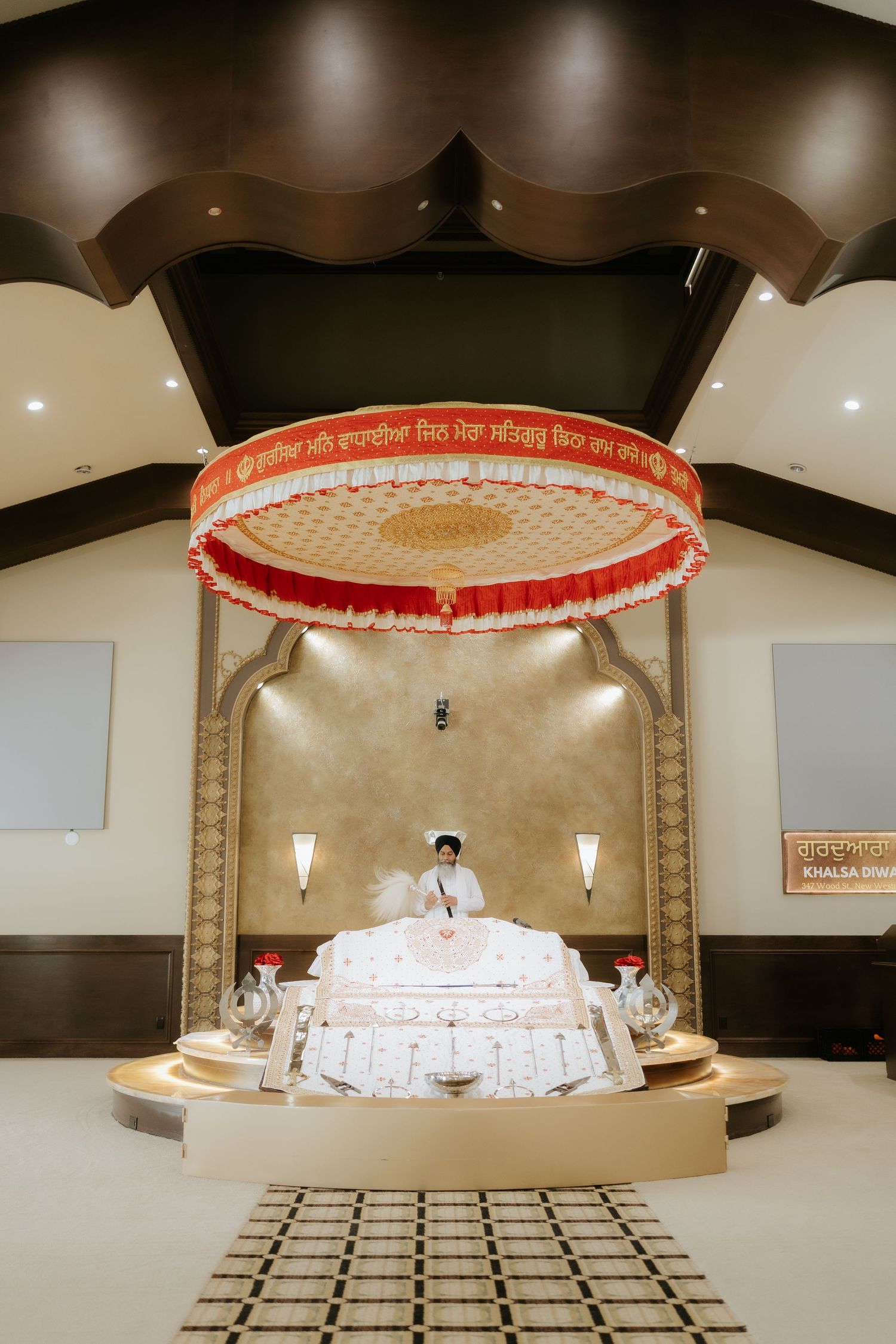 Ornate Sikh Gurdwara interior with Palki Sahib throne, decorated canopy, and Granthi seated during religious ceremony.