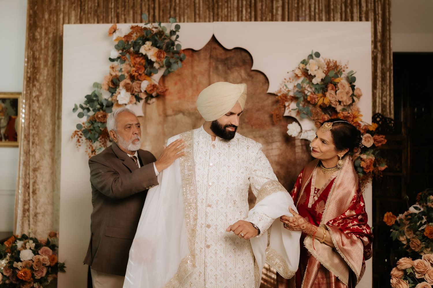 Sikh groom in white sherwani being dressed by parents before wedding ceremony with floral backdrop.