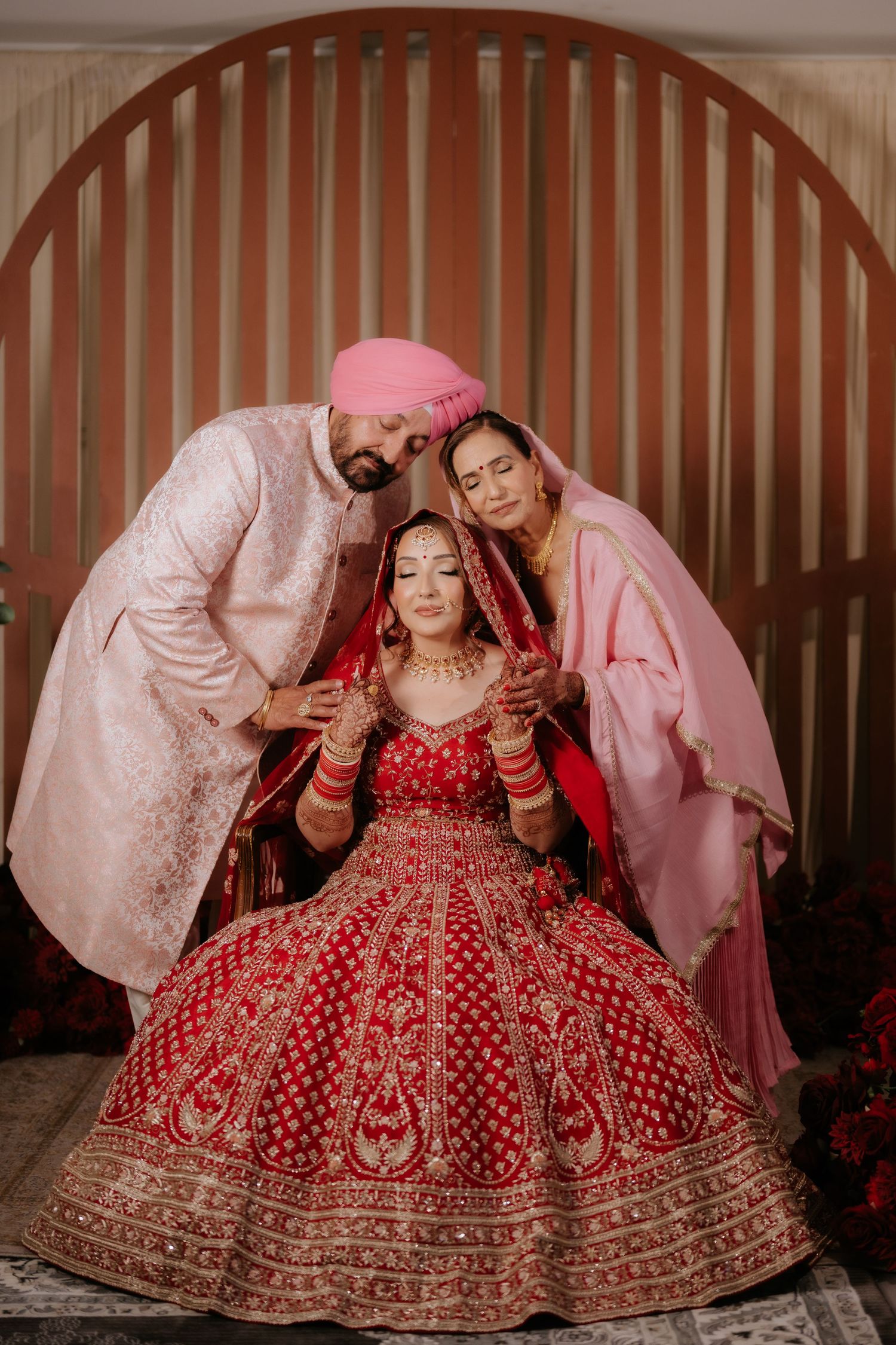 Indian bride in red lehenga seated with parents in traditional attire during Sikh wedding ceremony.