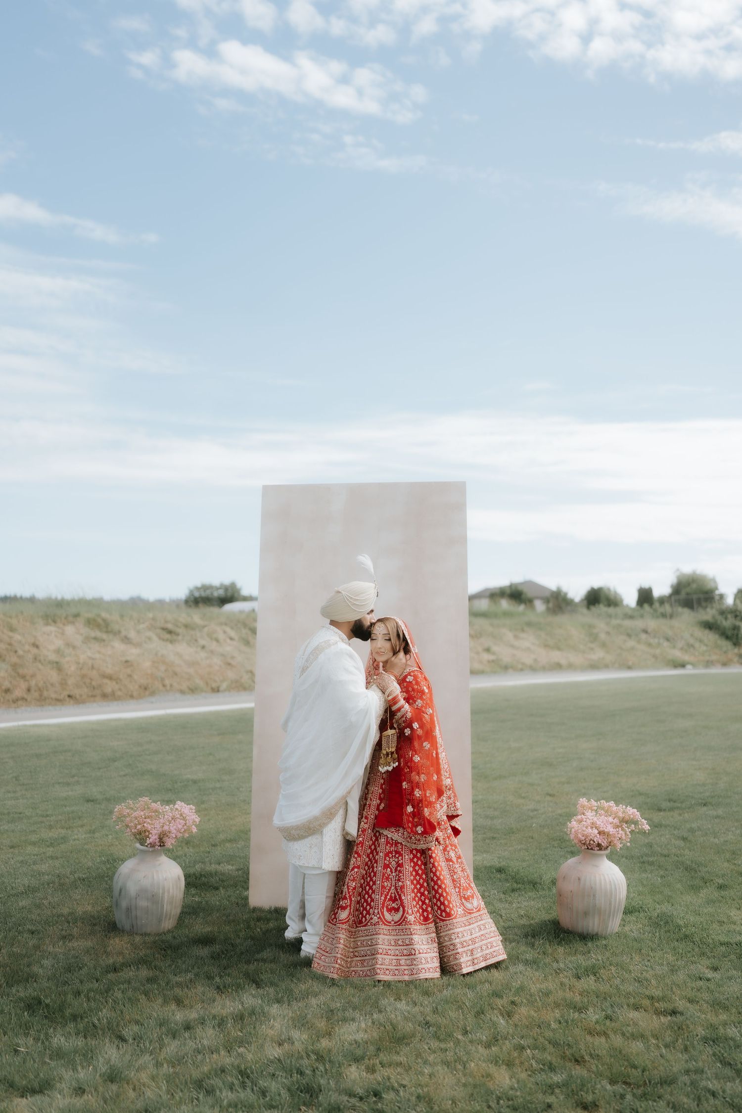 Indian bride in red lehenga and groom in white sherwani share an intimate moment at outdoor wedding ceremony.