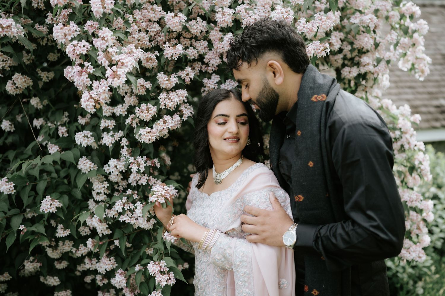 A couple poses romantically among blooming pink flowers in an outdoor garden setting.