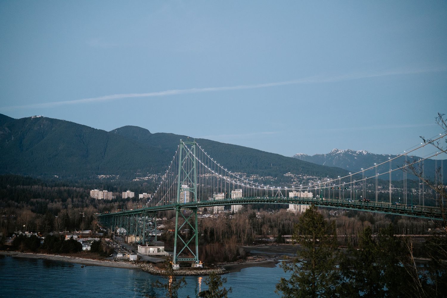 Lions Gate Bridge spans the Burrard Inlet with snow-capped mountains and city skyline in Vancouver, British Columbia.