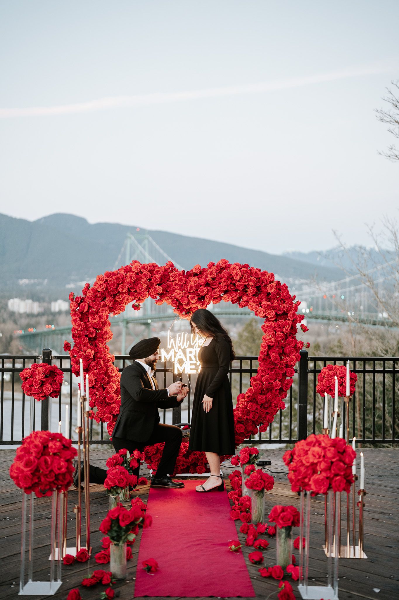 A romantic red floral arch installation with rose decorations along a pink carpet walkway overlooking a waterfront view.