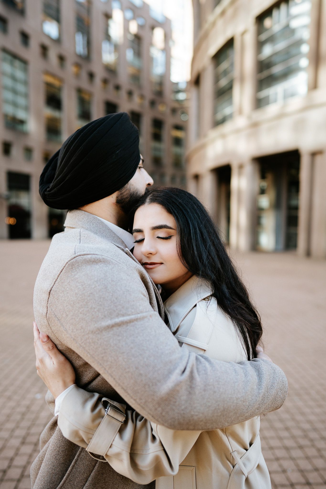 A couple shares an intimate embrace in front of a modern urban building with stone architecture.