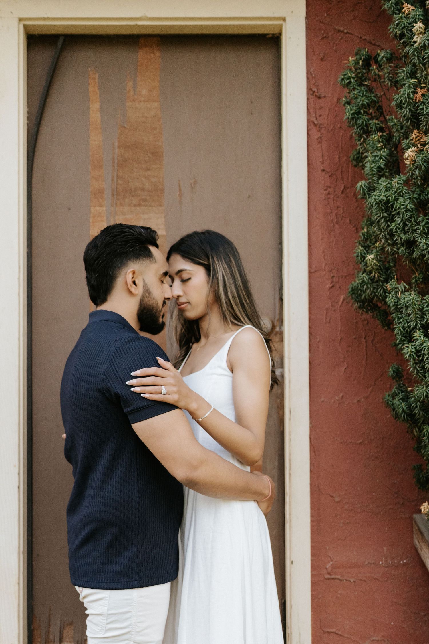 A romantic couple embraces in a doorway against a terracotta wall with climbing vines.