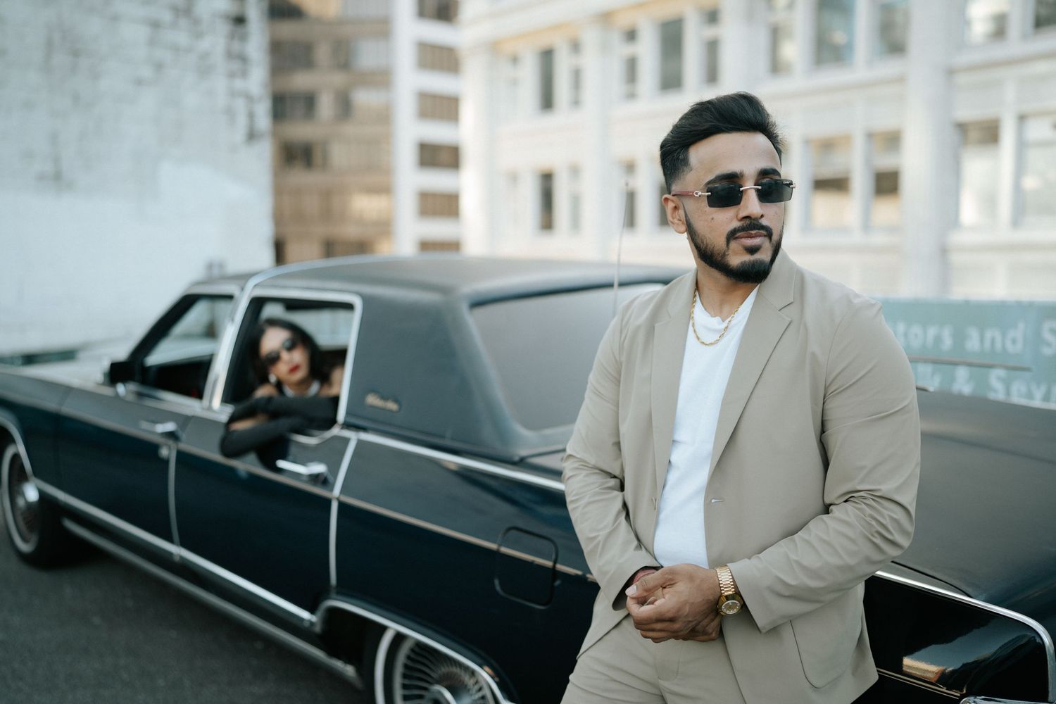 A person in a beige suit leans against a sleek black vintage car on a city street.