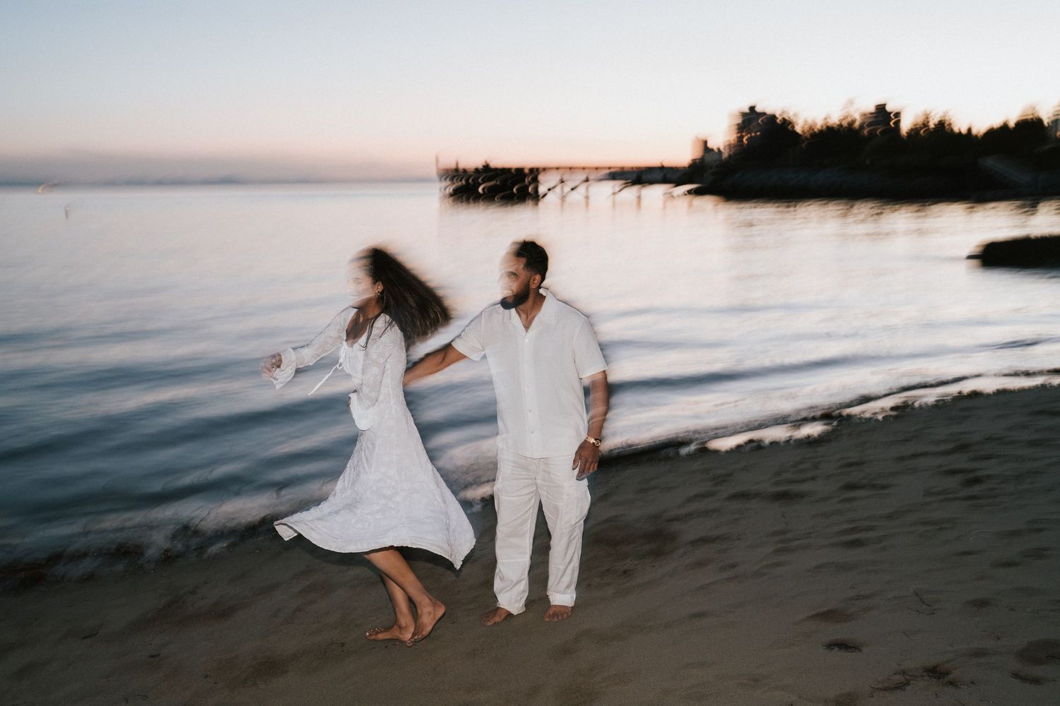 Couple in white outfits walking along a beach at sunset creating silhouettes against the ocean backdrop.