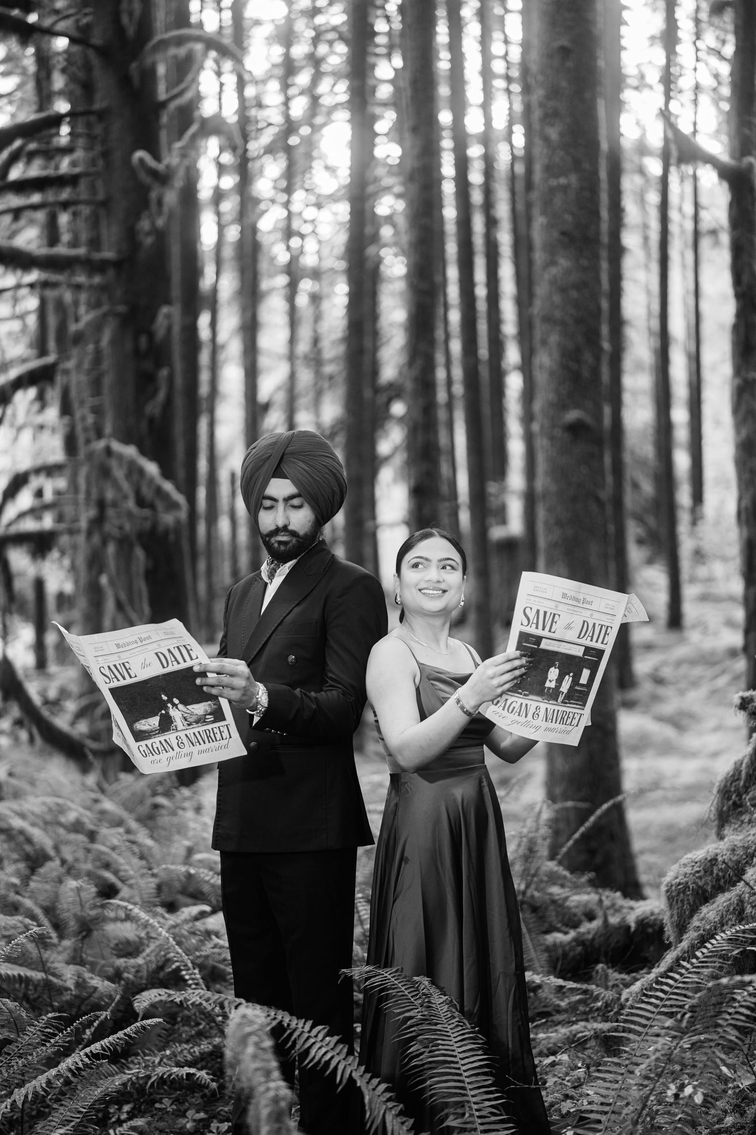 Couple poses with newspapers for engagement photos surrounded by tall trees and ferns.