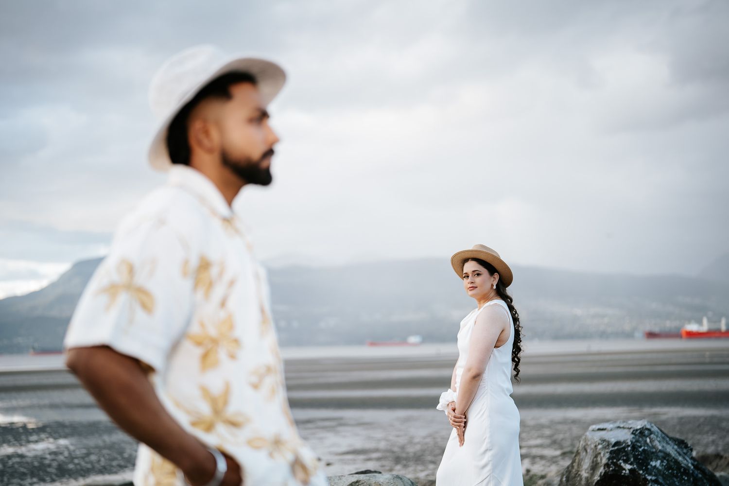 A woman in a white dress and straw hat gazes back at a bearded man in a floral shirt on a beach.