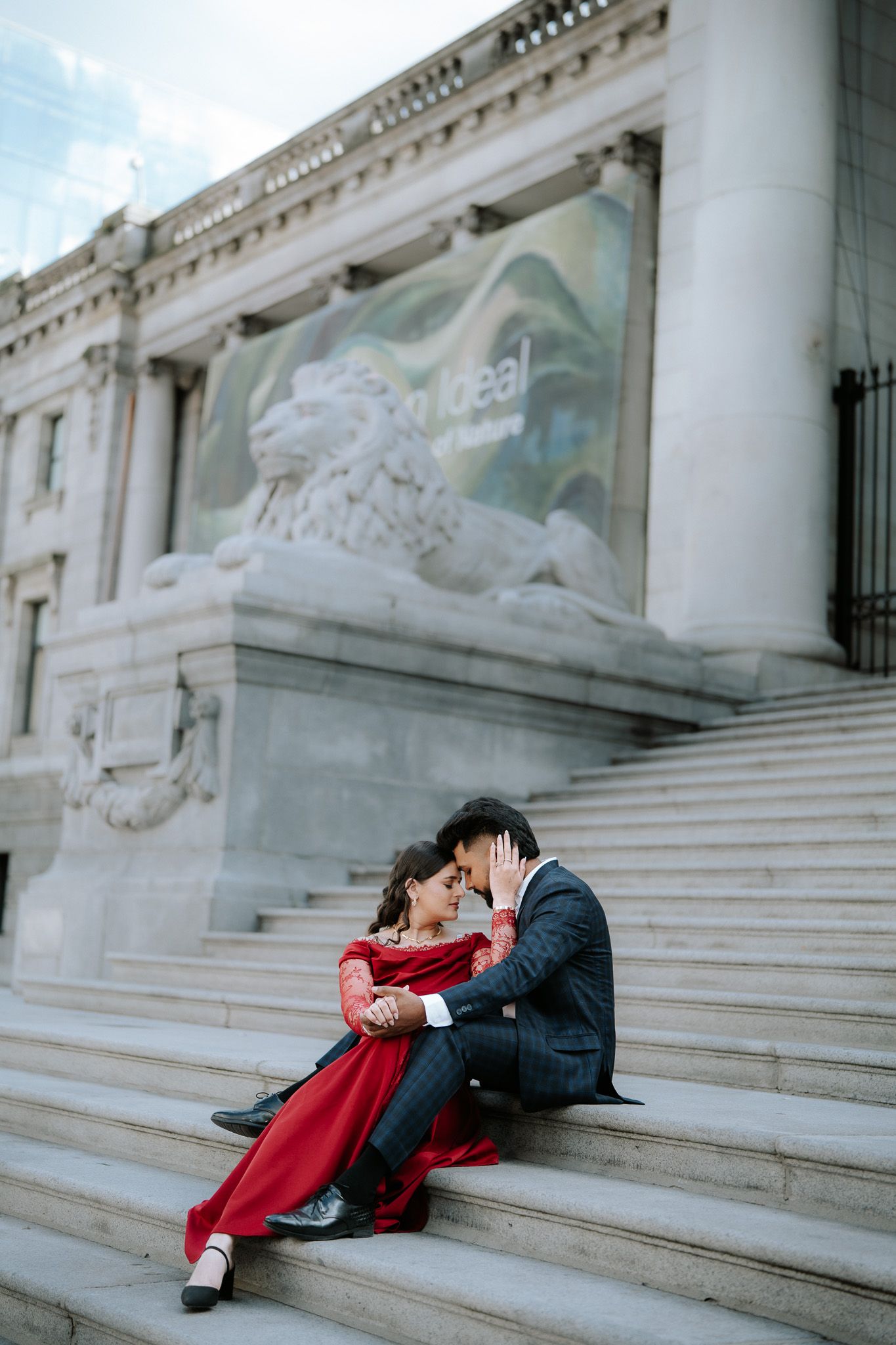Couple in red dress and suit sitting intimately on steps of a grand neoclassical building with lion sculpture.