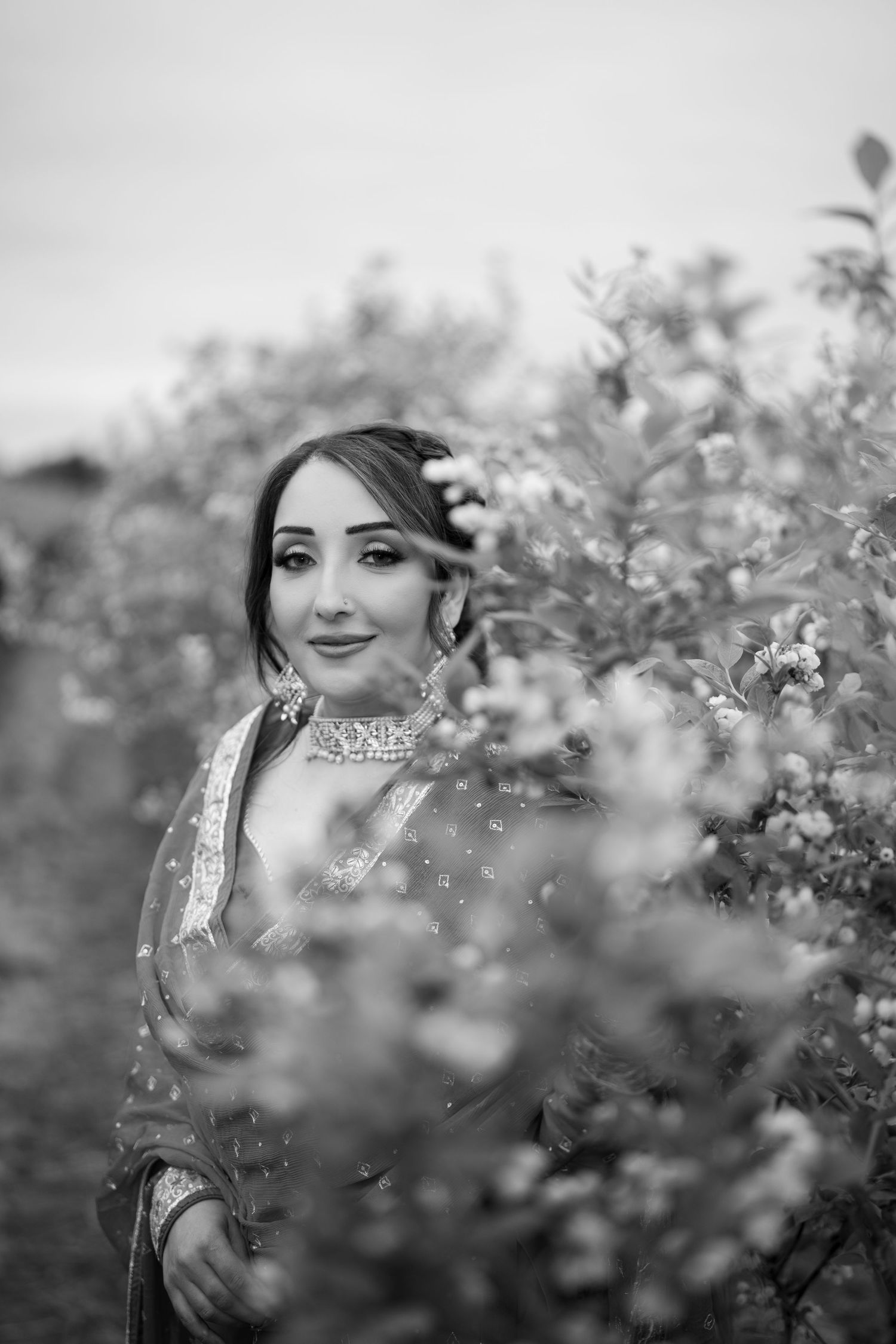 Black and white portrait of a bride in traditional South Asian attire surrounded by blooming flowers.