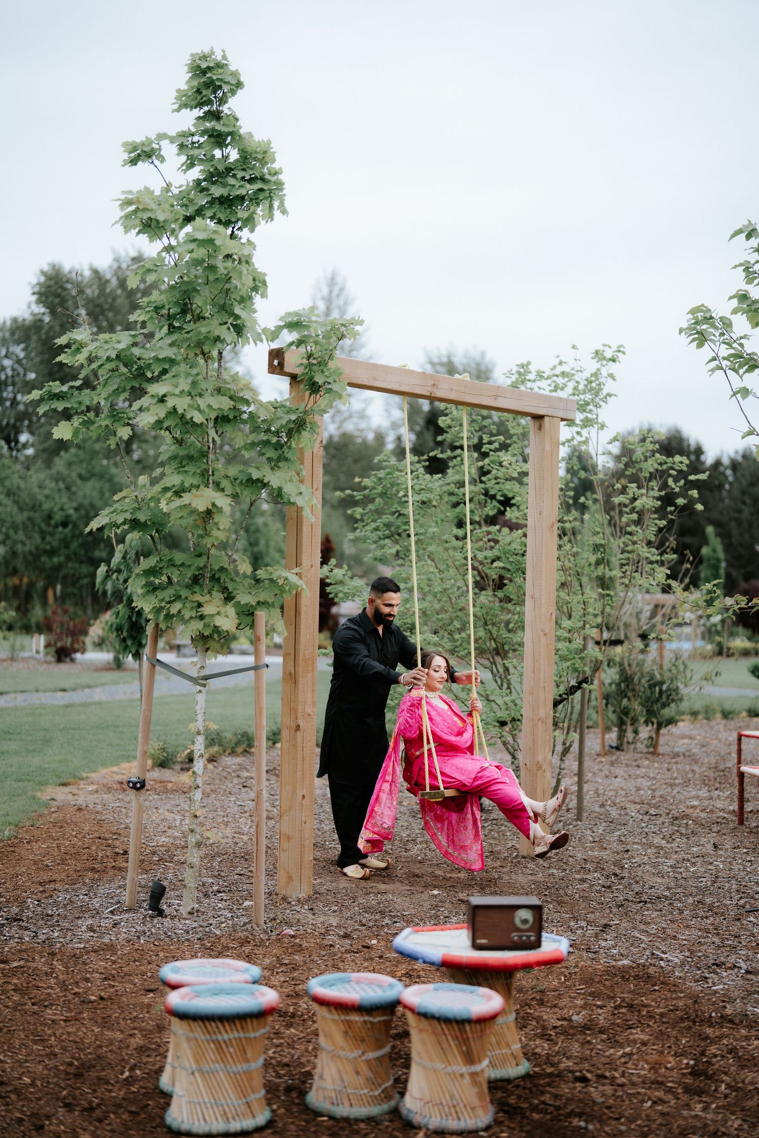 Bride in pink lehenga on wooden swing pushed by groom in black sherwani at outdoor garden wedding venue.