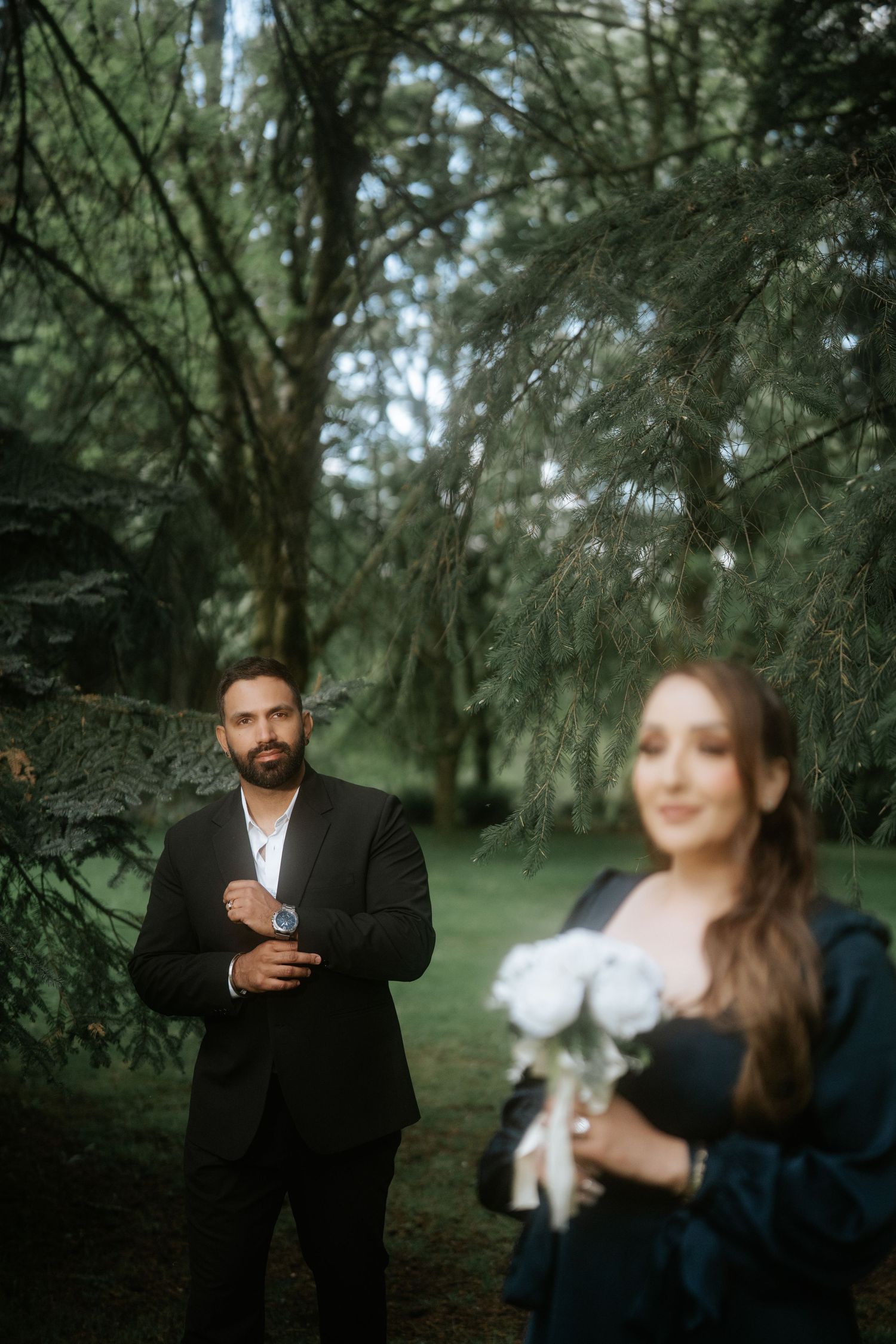 A bearded groom in a black suit adjusts his cufflinks while a bride holding white roses stands in soft focus outdoors.