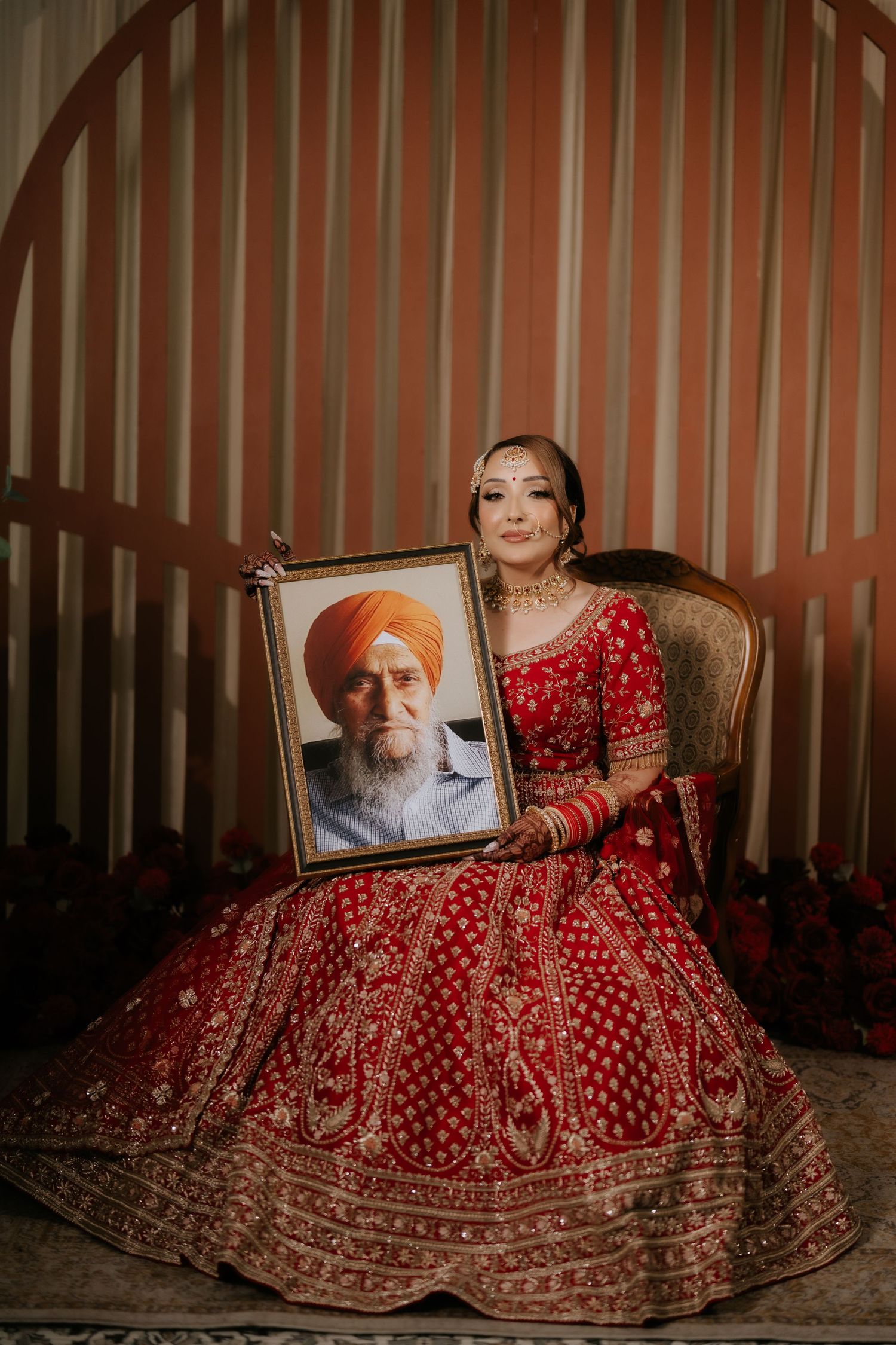 Sikh bride in red lehenga holding framed portrait of elderly man wearing orange turban, seated on ornate chair.
